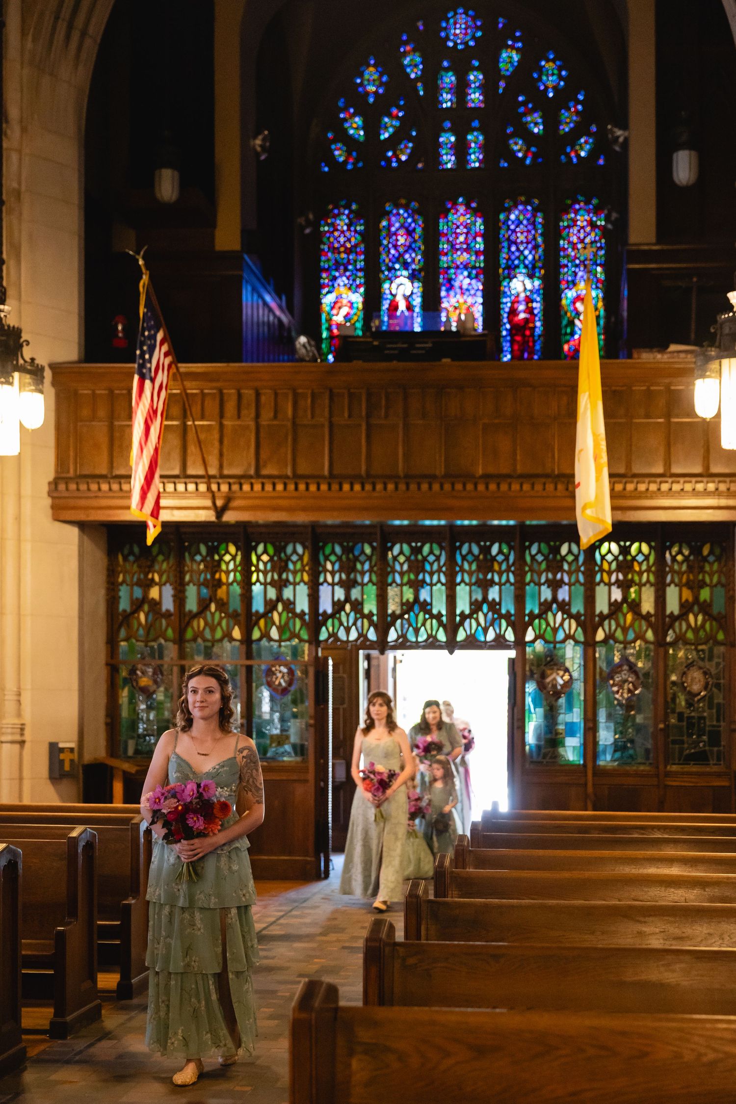 Wedding party walks down church aisle with stained glass windows casting colorful light throughout the sanctuary.