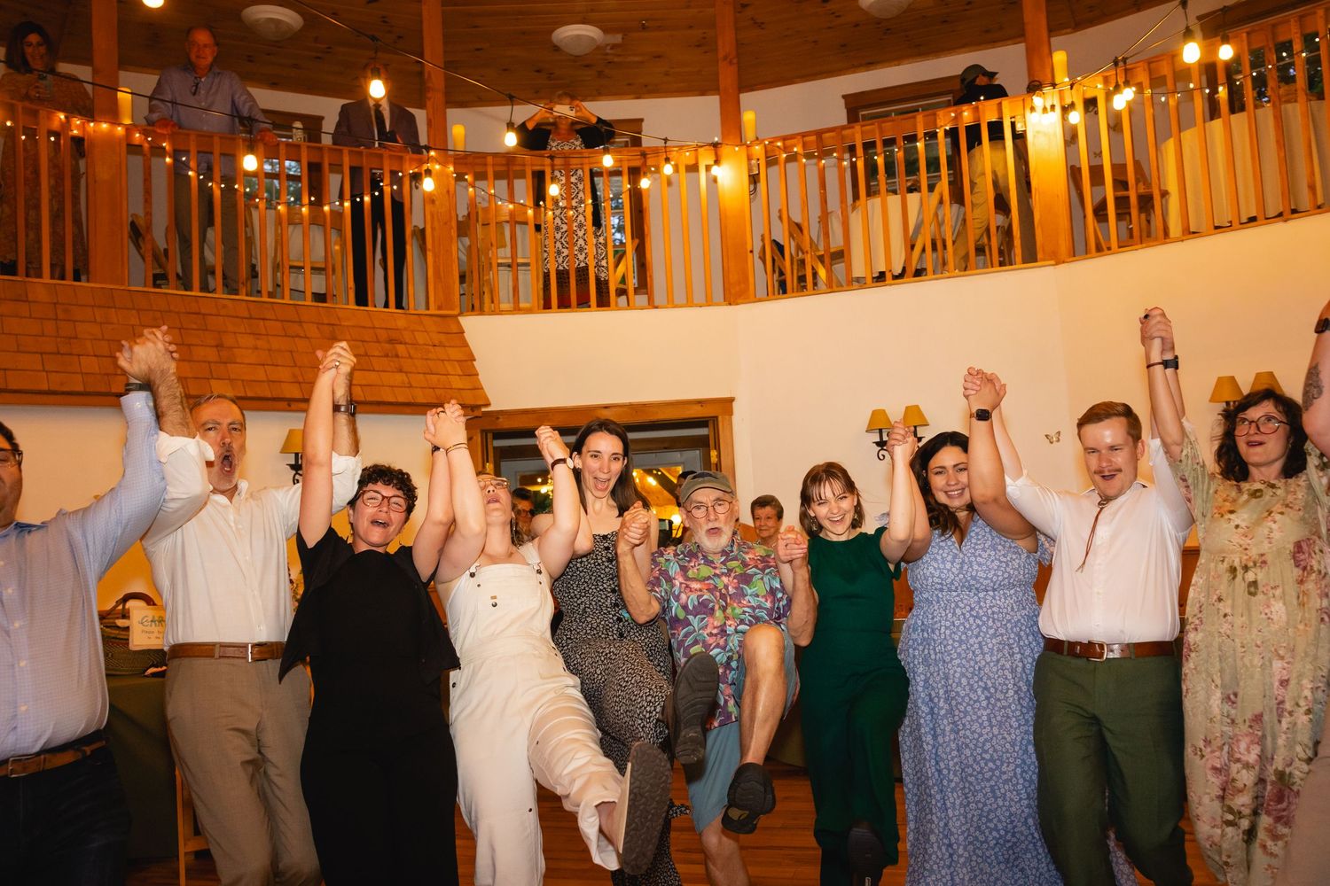 A group of wedding guests dancing and celebrating with raised arms under warm lighting in a rustic wooden venue.