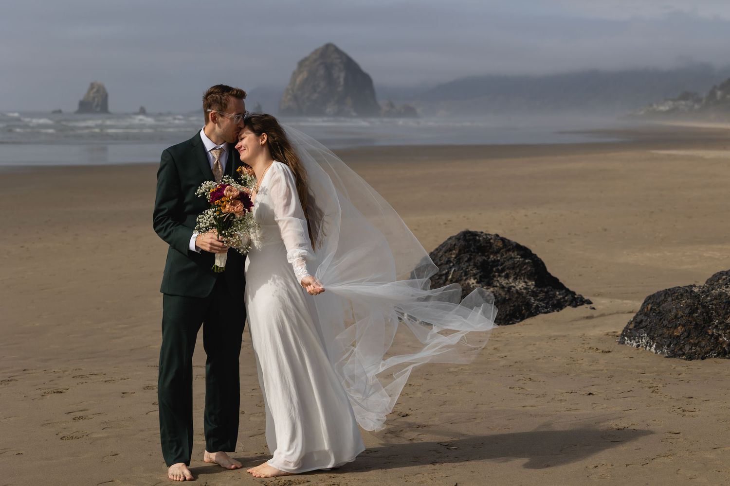 A couple shares a romantic moment on a misty beach with Haystack Rock visible in the background at Cannon Beach, Oregon.