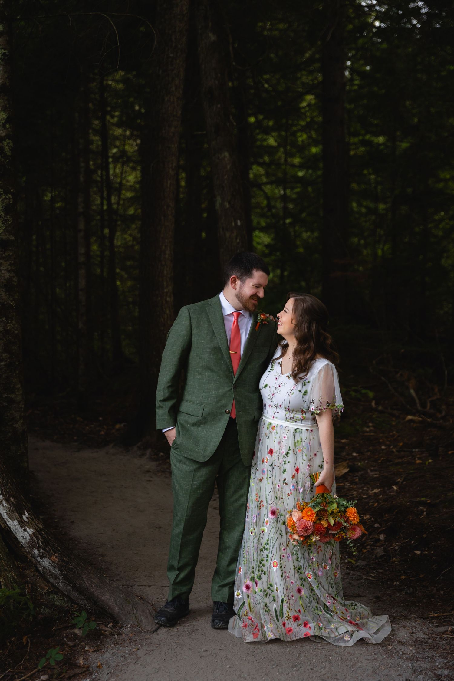 A couple in wedding attire stands on a forest path, with the bride holding a vibrant orange and red bouquet.
