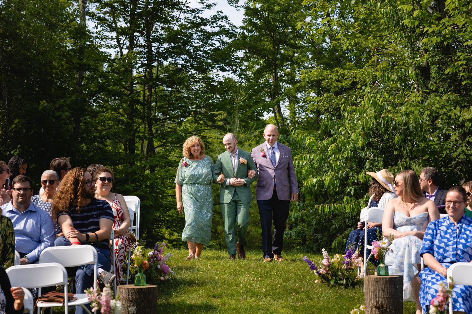 Guests gather outdoors for a sunny summer wedding ceremony in a lush green garden setting.