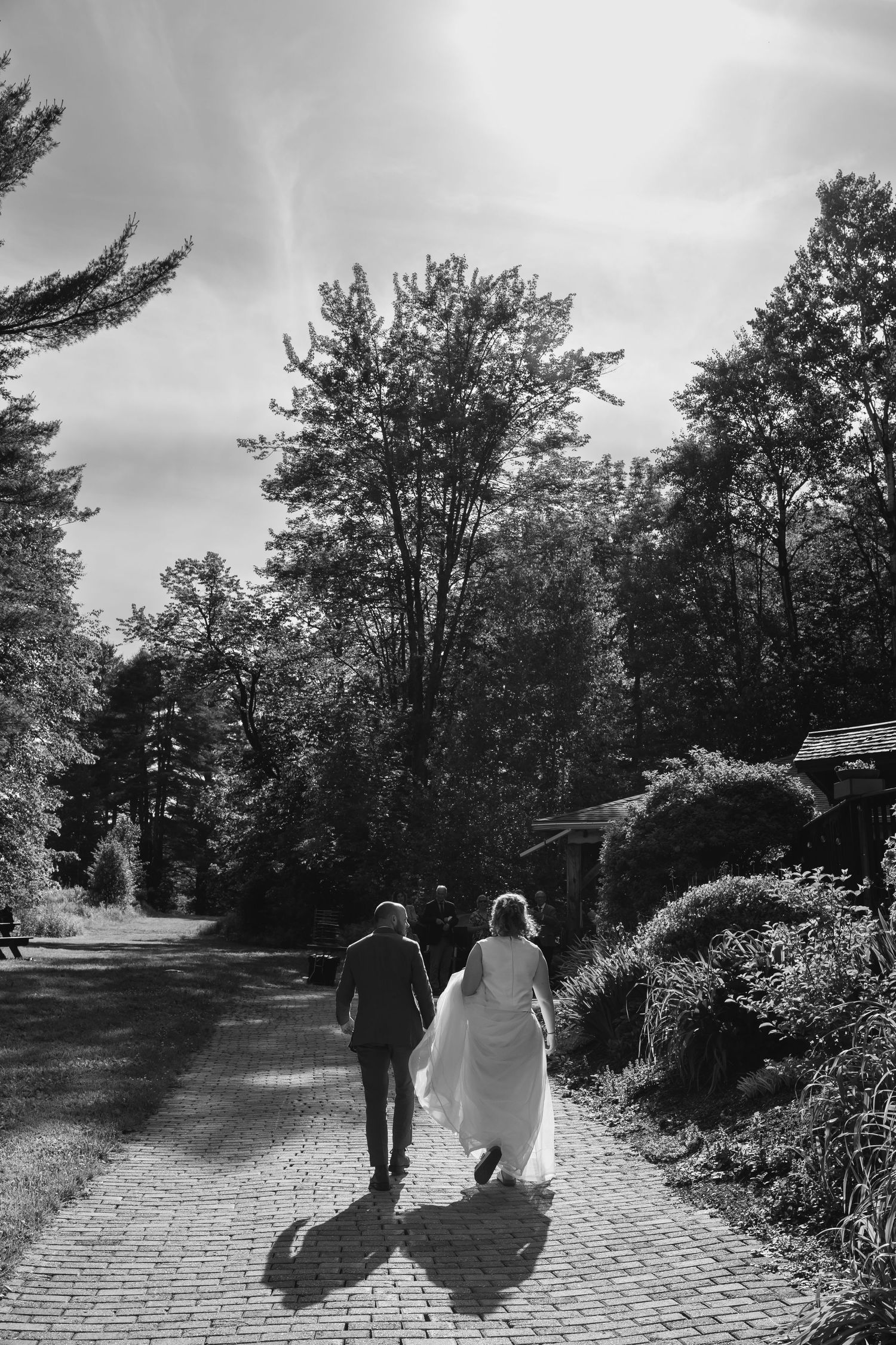 Two people walking together on a winding brick path through a scenic wooded area on a sunny day.