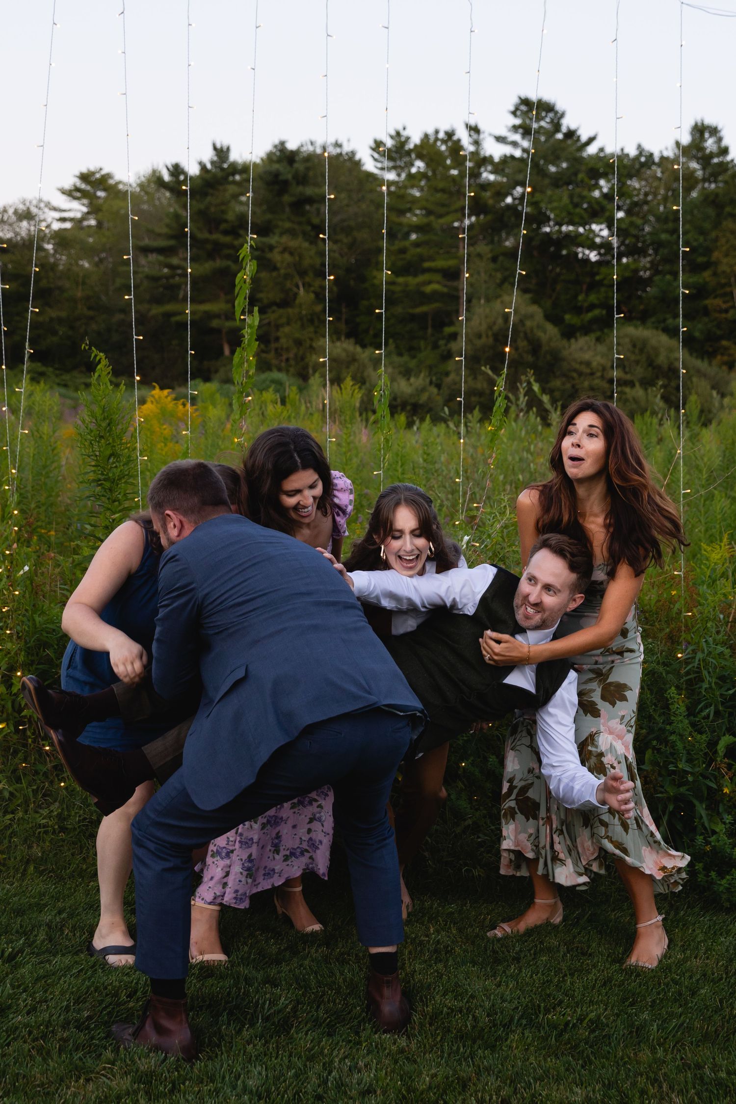 A group of people playfully falling over in sequence on a grassy field with pine trees in the background.