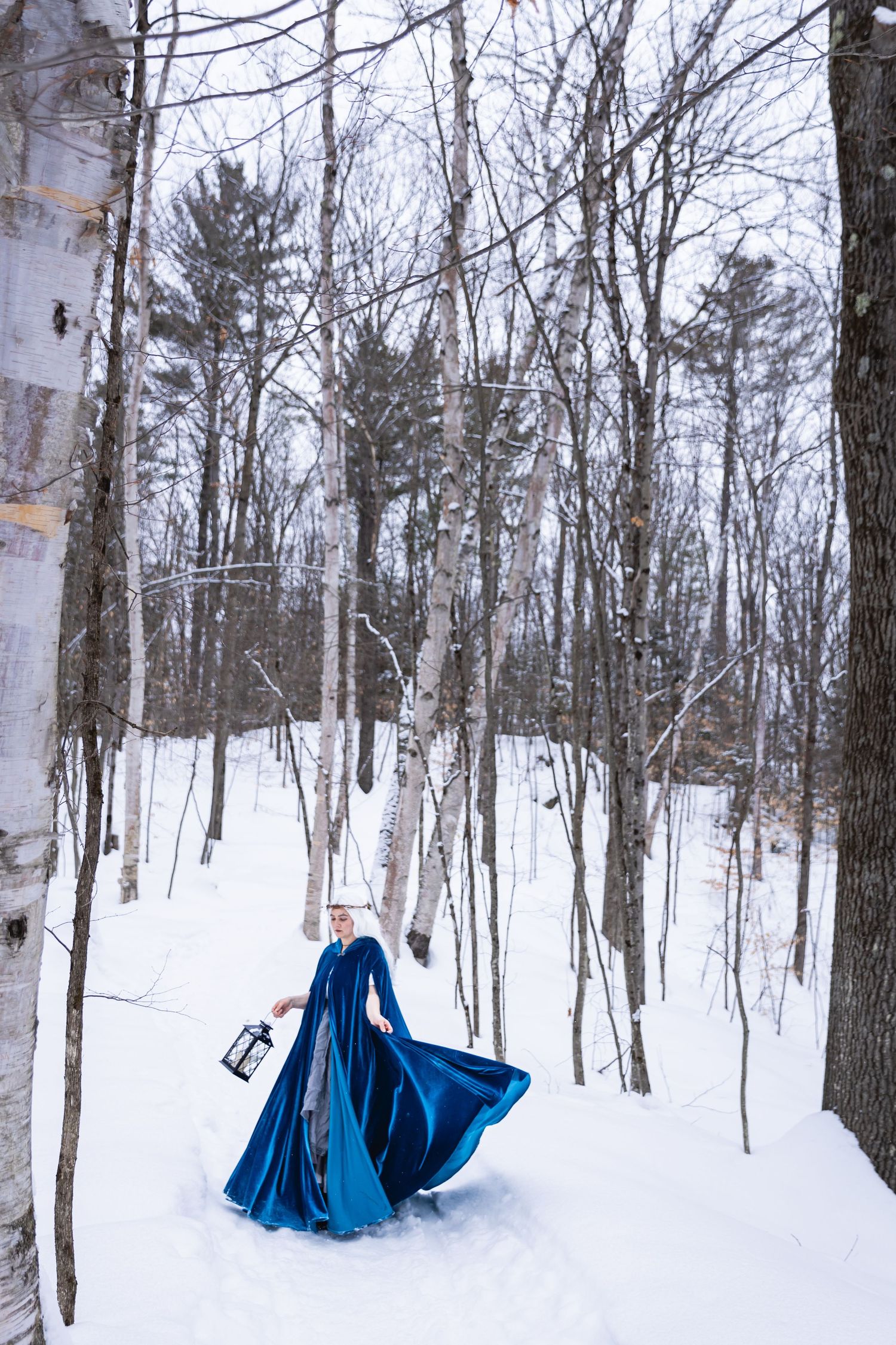 Woman in flowing blue velvet cloak walking through snowy winter forest.