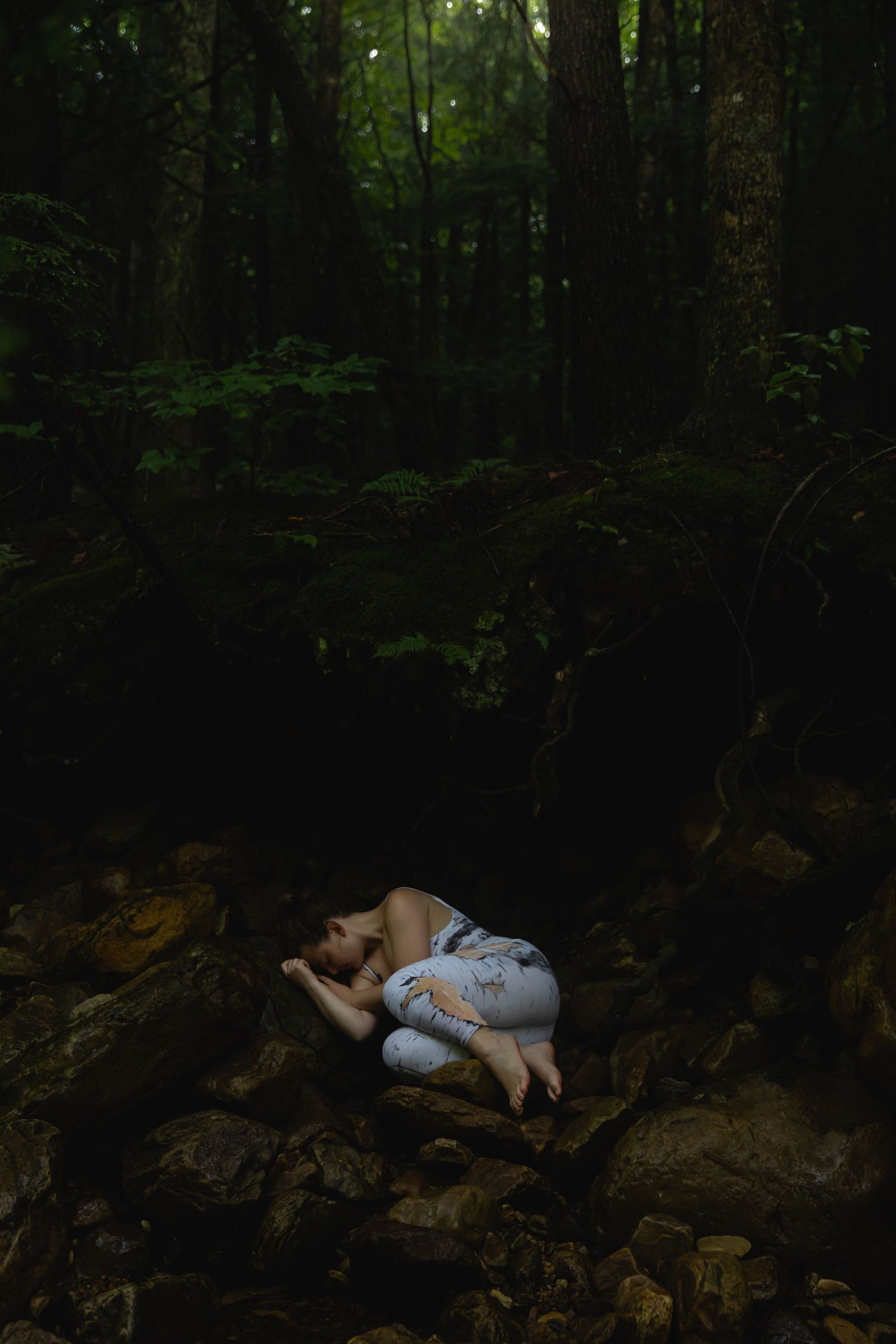 Woman in white dress curled up sleeping on rocks in a dark lush green forest.