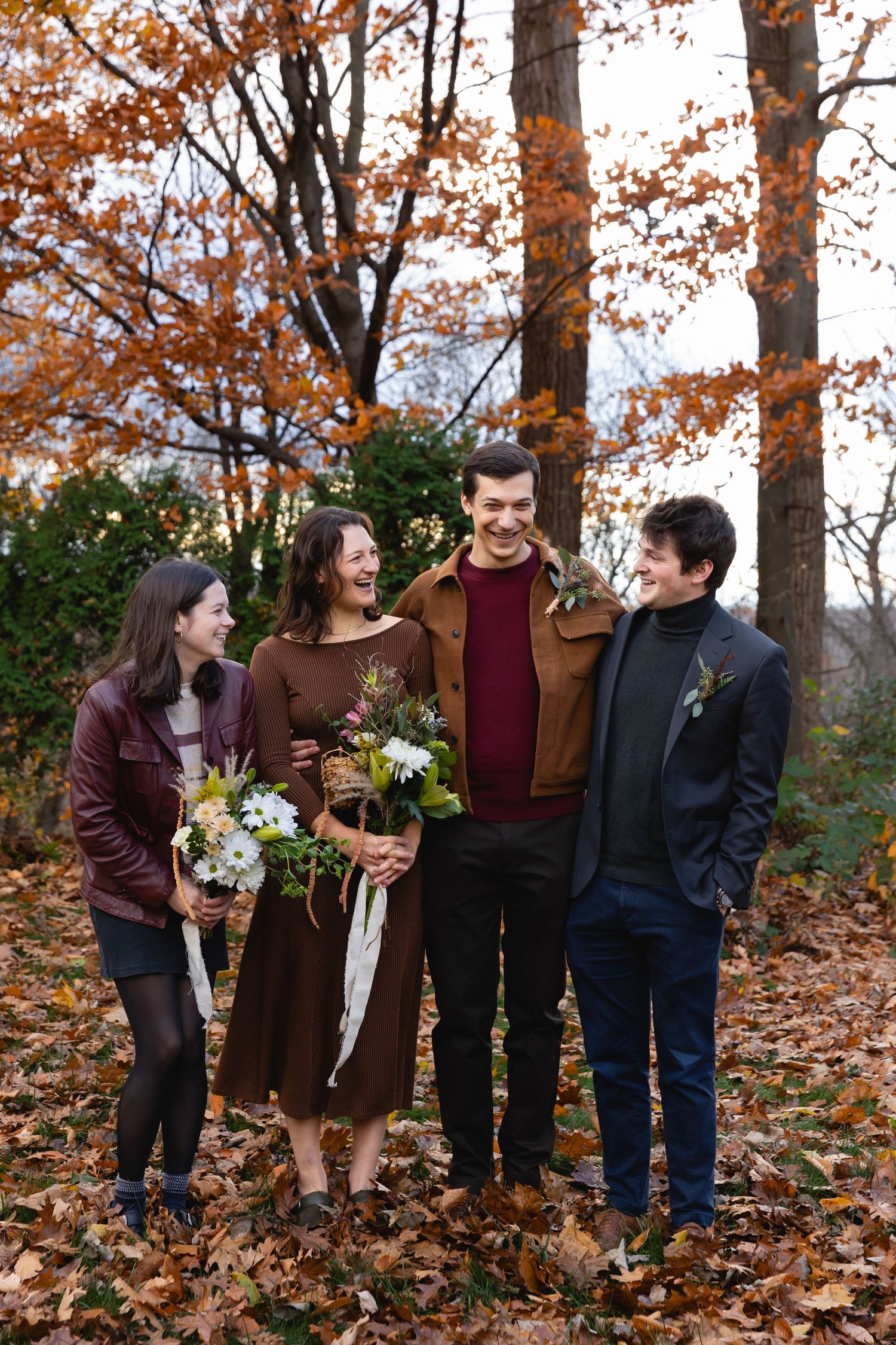 A group stands together laughing in a fall forest setting with autumn leaves and bouquets of flowers.