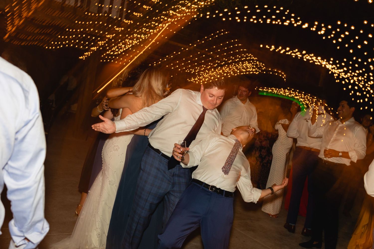 People dancing at a wedding reception under twinkly string lights in a barn venue.