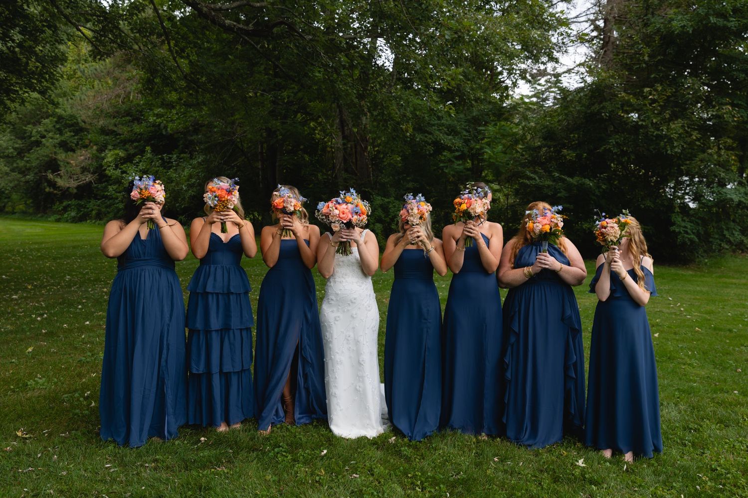 Bridesmaids in matching navy blue dresses pose with bride in white gown on grassy lawn while covering their faces.