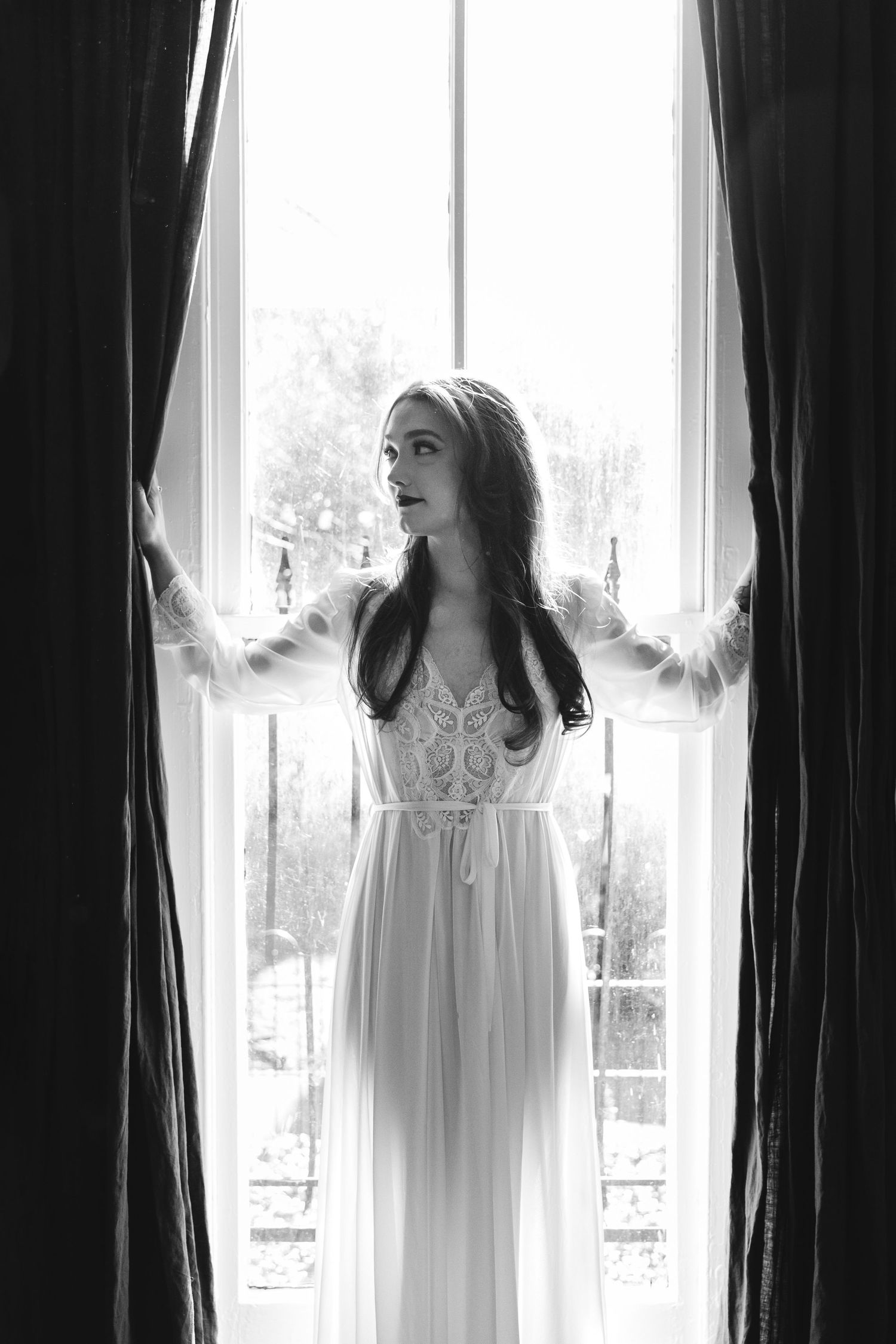 Black and white photo of a woman in a white lace dress standing between curtains by a bright window.