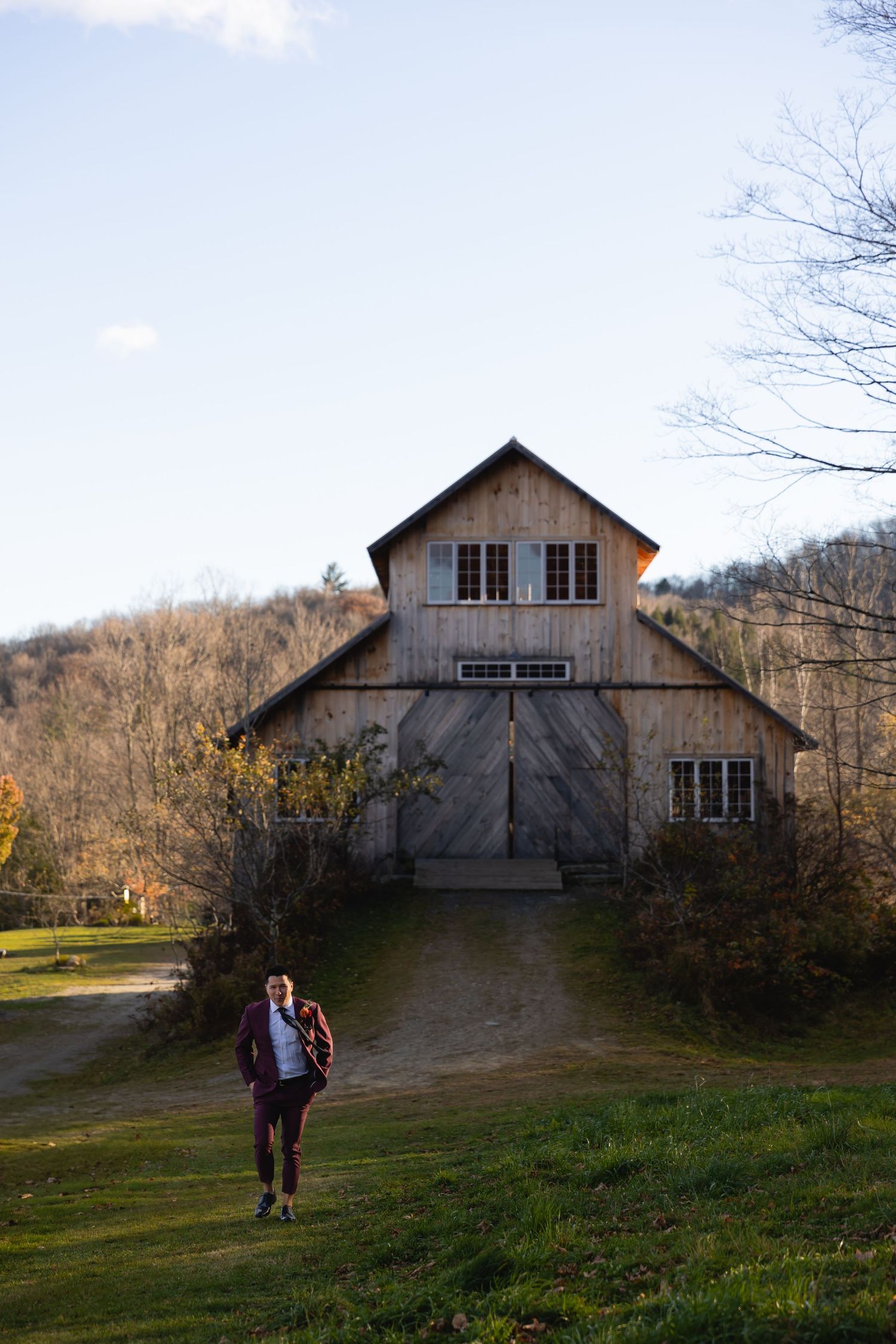 A person walks on a grassy path in front of a rustic wooden barn with large doors in an autumn rural setting.