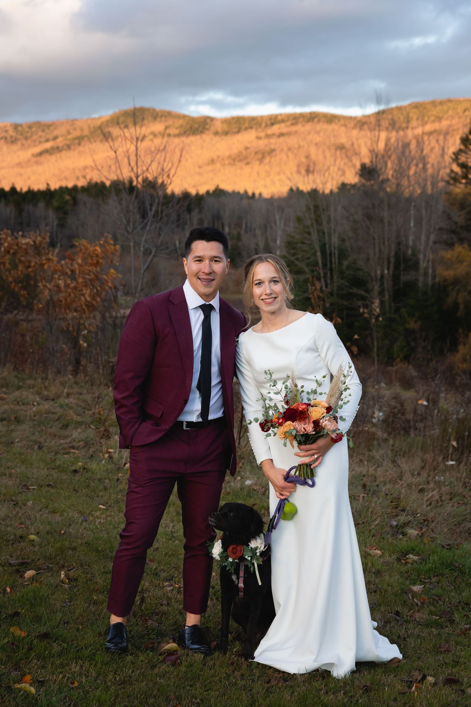 A couple poses in wedding attire in front of autumn mountains at sunset, wearing burgundy suit and white gown with flowers.