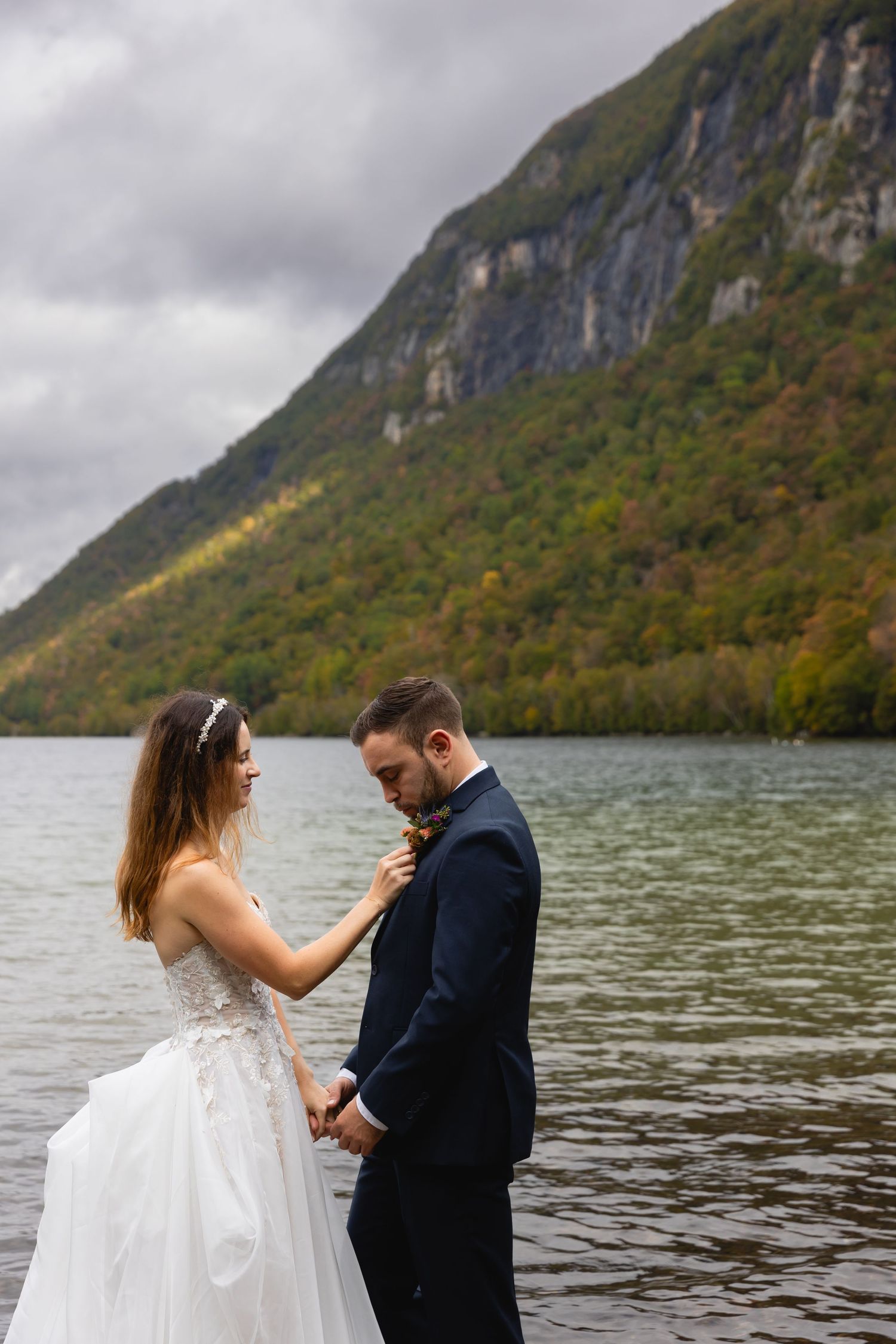 Couple in wedding attire standing by mountain lake holding hands intimately.