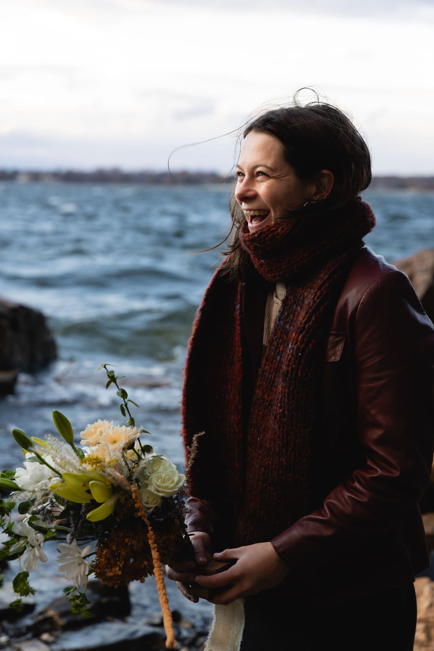 A joyful moment captured by the ocean holding a bouquet of yellow and white flowers while wearing a brown leather jacket.