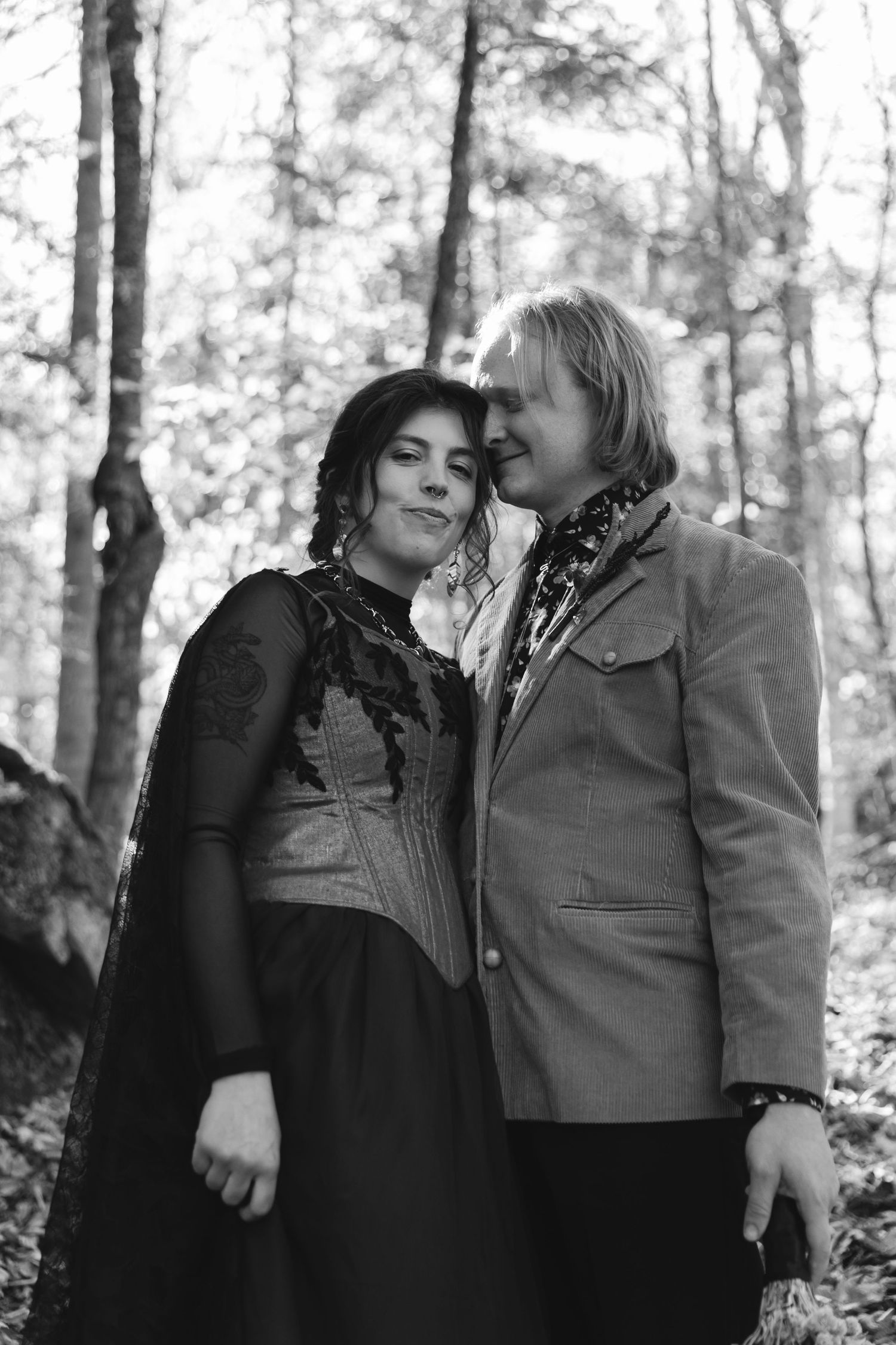 Black and white photo of couple embracing in a forest, woman in black lace dress leaning on man.