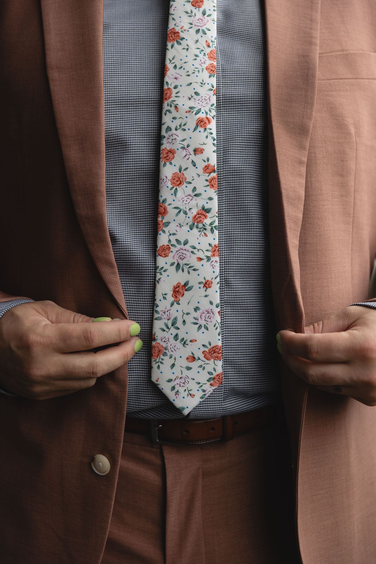 Groom in pink suit adjusting floral tie with green manicured nails visible.