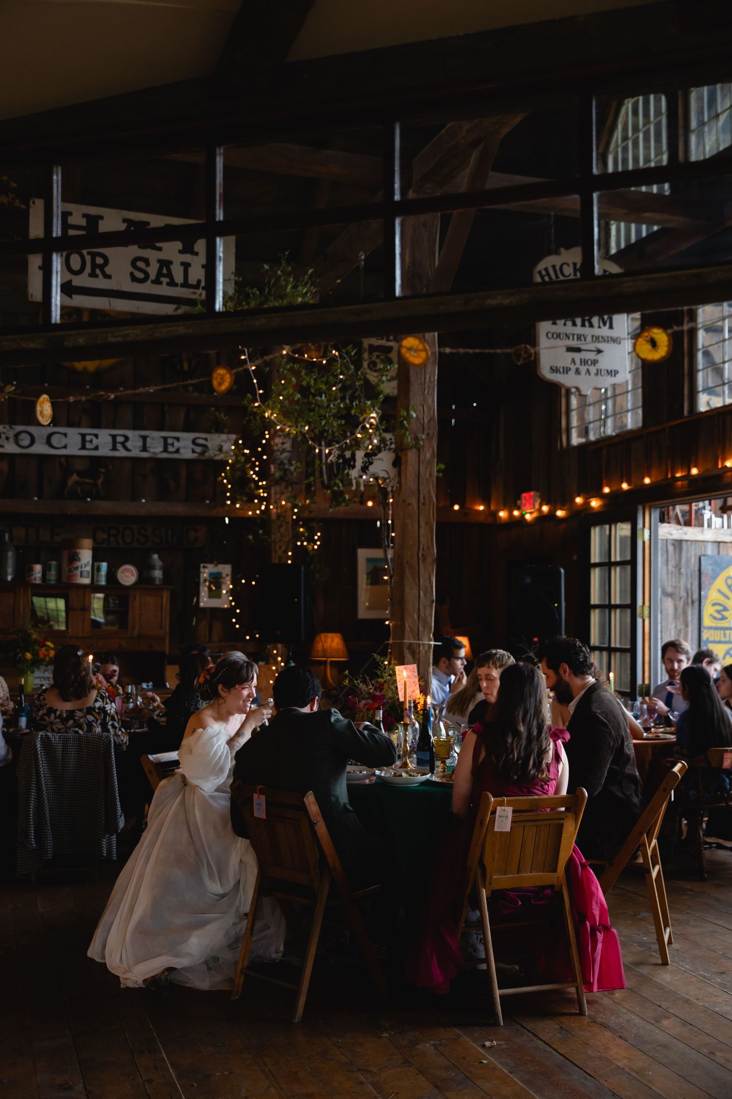 Wedding reception in a rustic barn venue with string lights, guests dining at candlelit tables in warm ambiance.