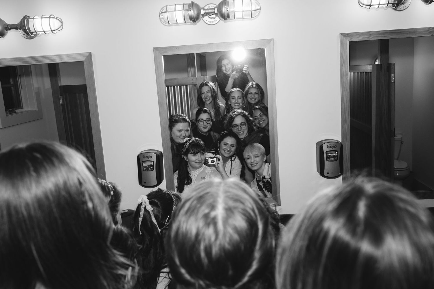 Black and white photo of bridal party crowding into bathroom mirror for group selfie.