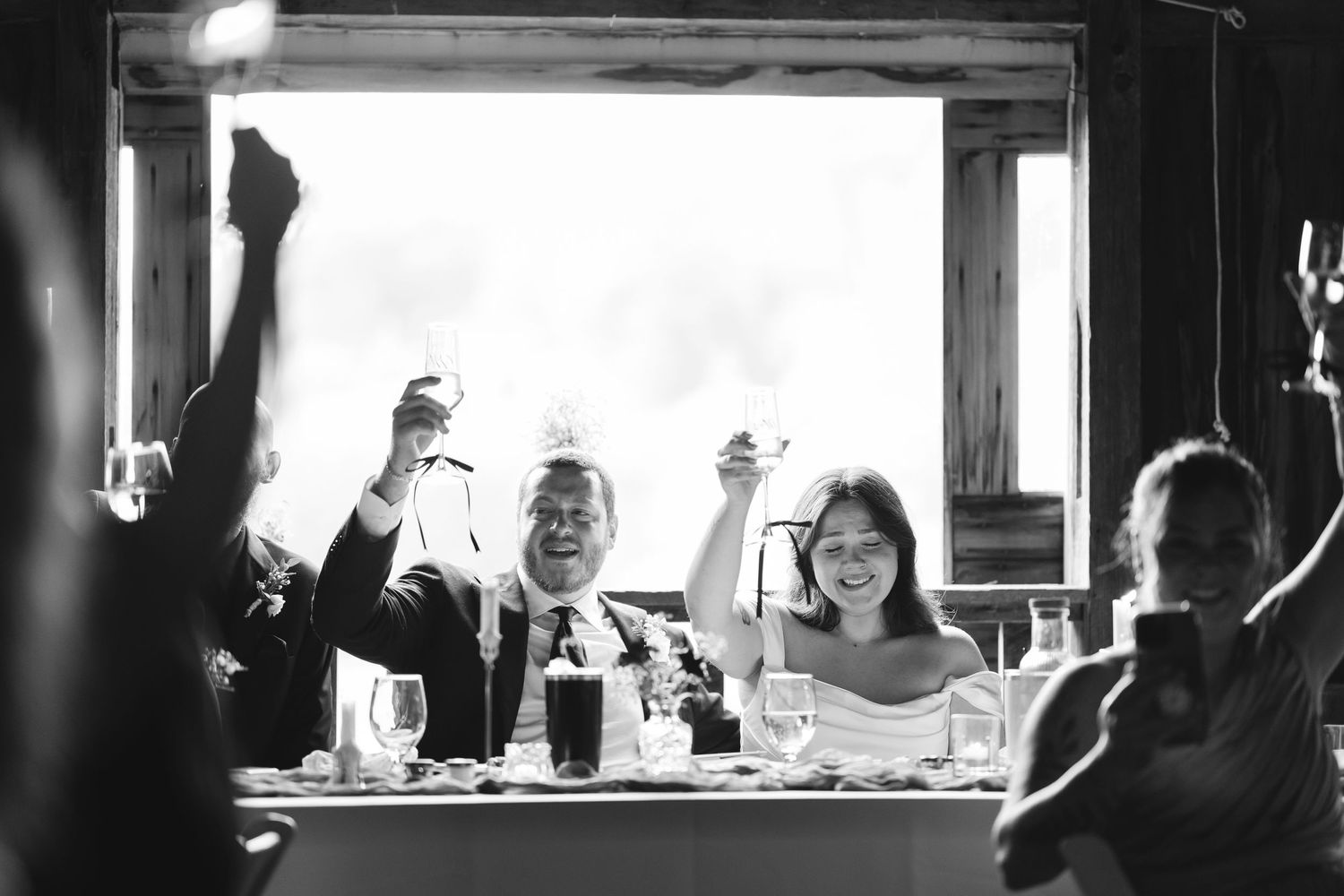 Bride and groom raise glasses in joyful toast with guests in black and white wedding reception photo.