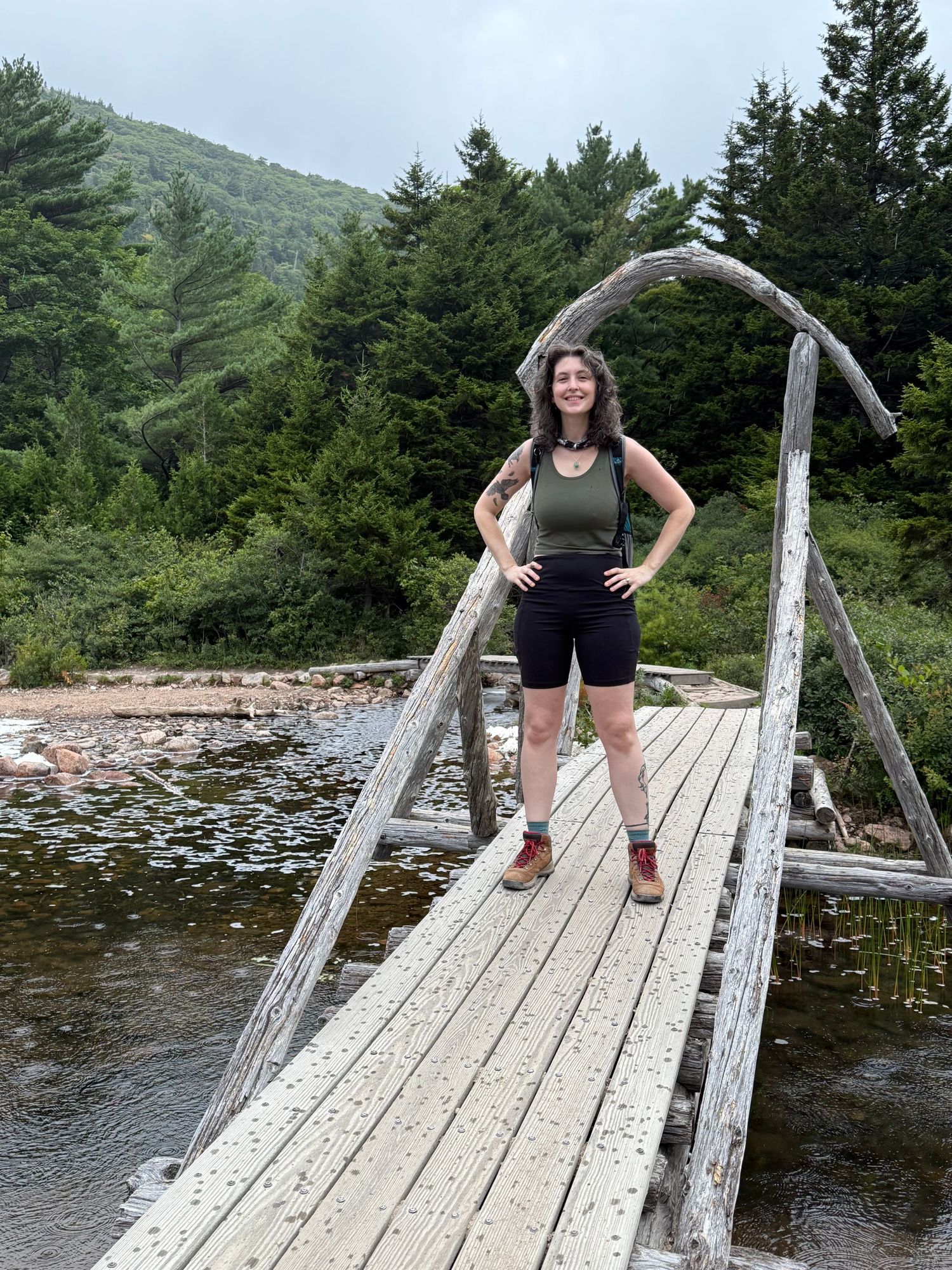Smiling woman in hiking gear standing on a rustic wooden suspension bridge in a mountain forest.