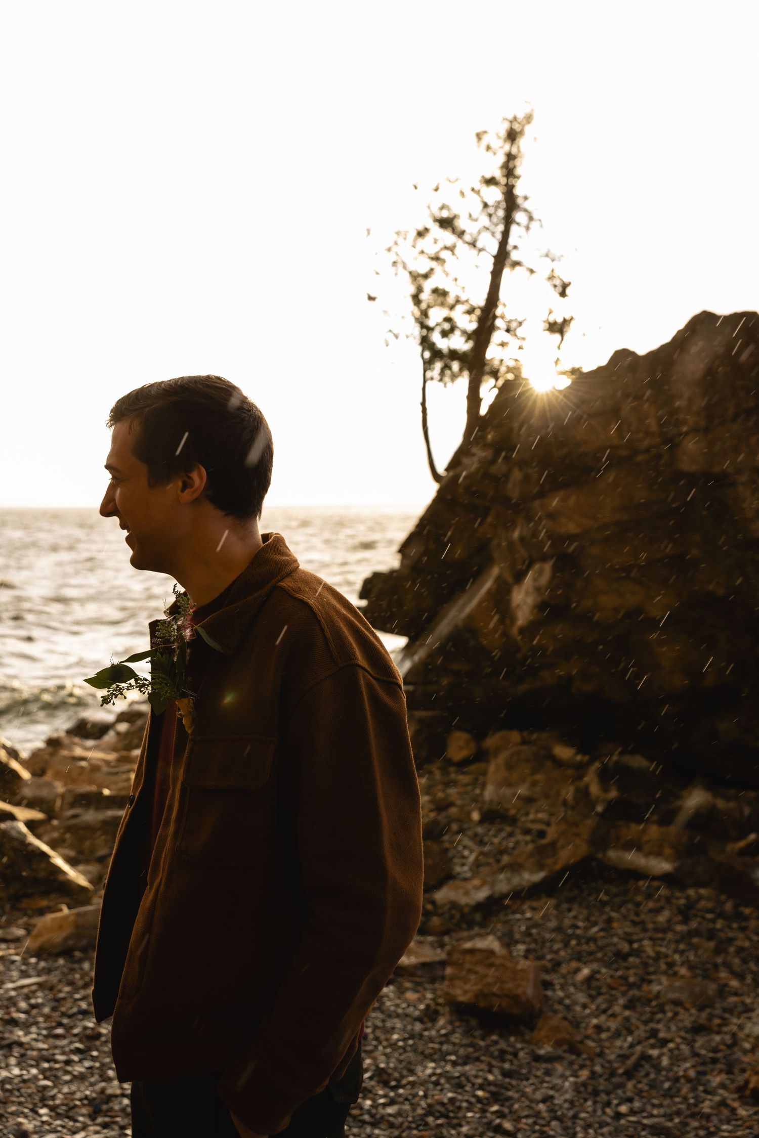 Man in brown jacket standing on rocky shoreline at golden sunset with ocean waves in background.