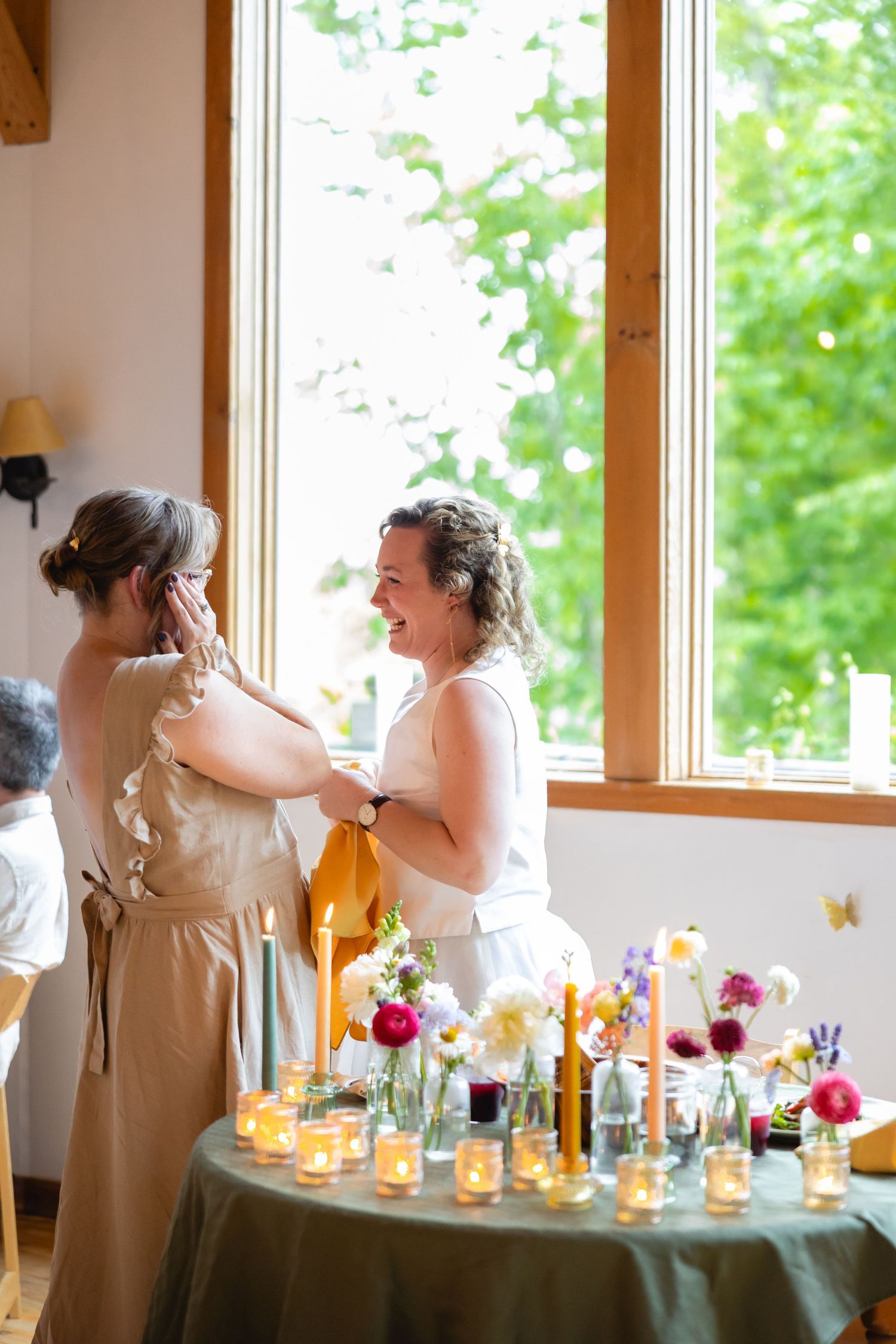Wedding reception table decorated with colorful flowers and glowing candles in glass holders against bright windows.