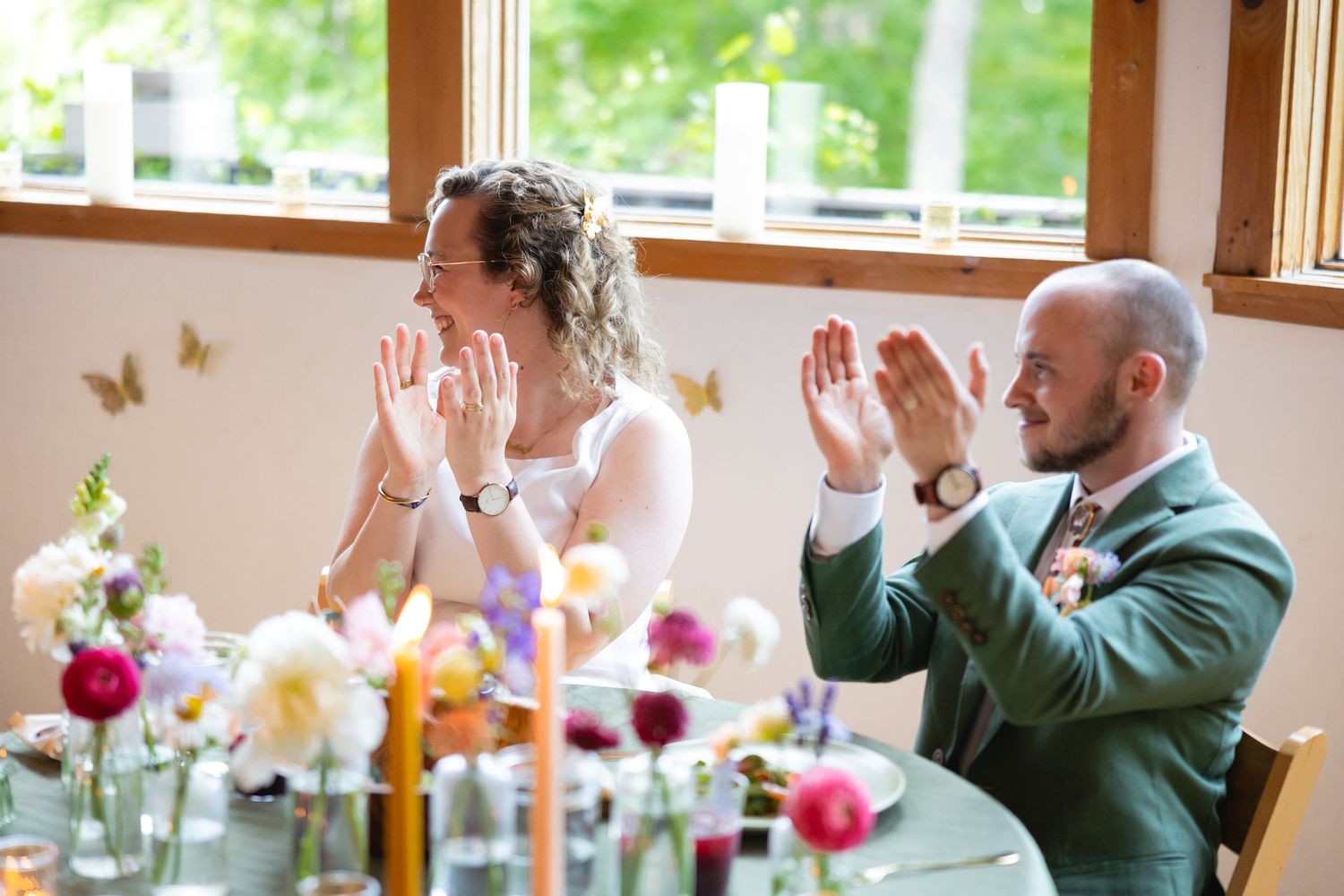 Wedding guests clap and celebrate during a reception in a bright, window-lit venue with colorful floral centerpieces.