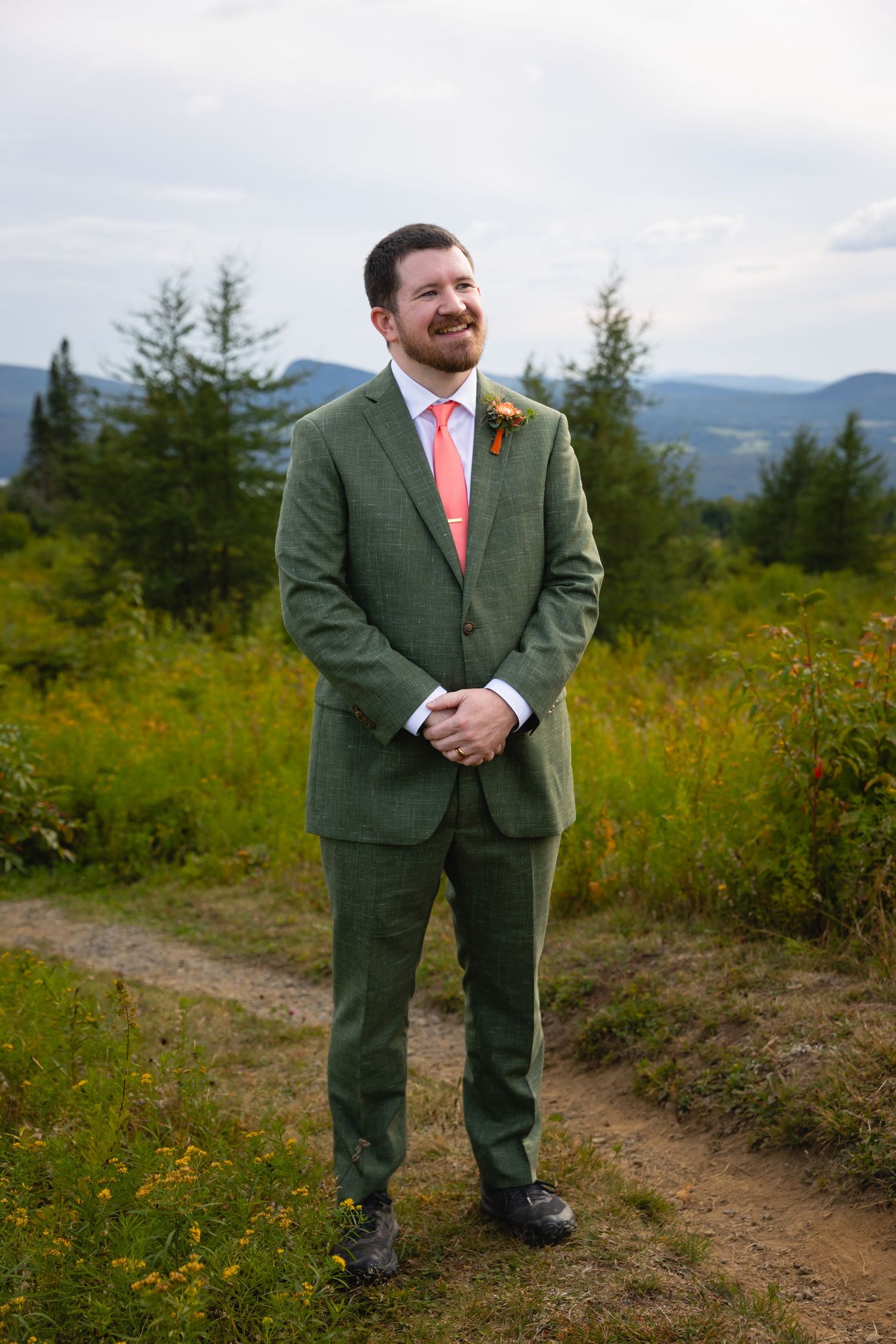 A person in a green suit and coral tie stands on a mountain trail with evergreen trees and mountains in the background.
