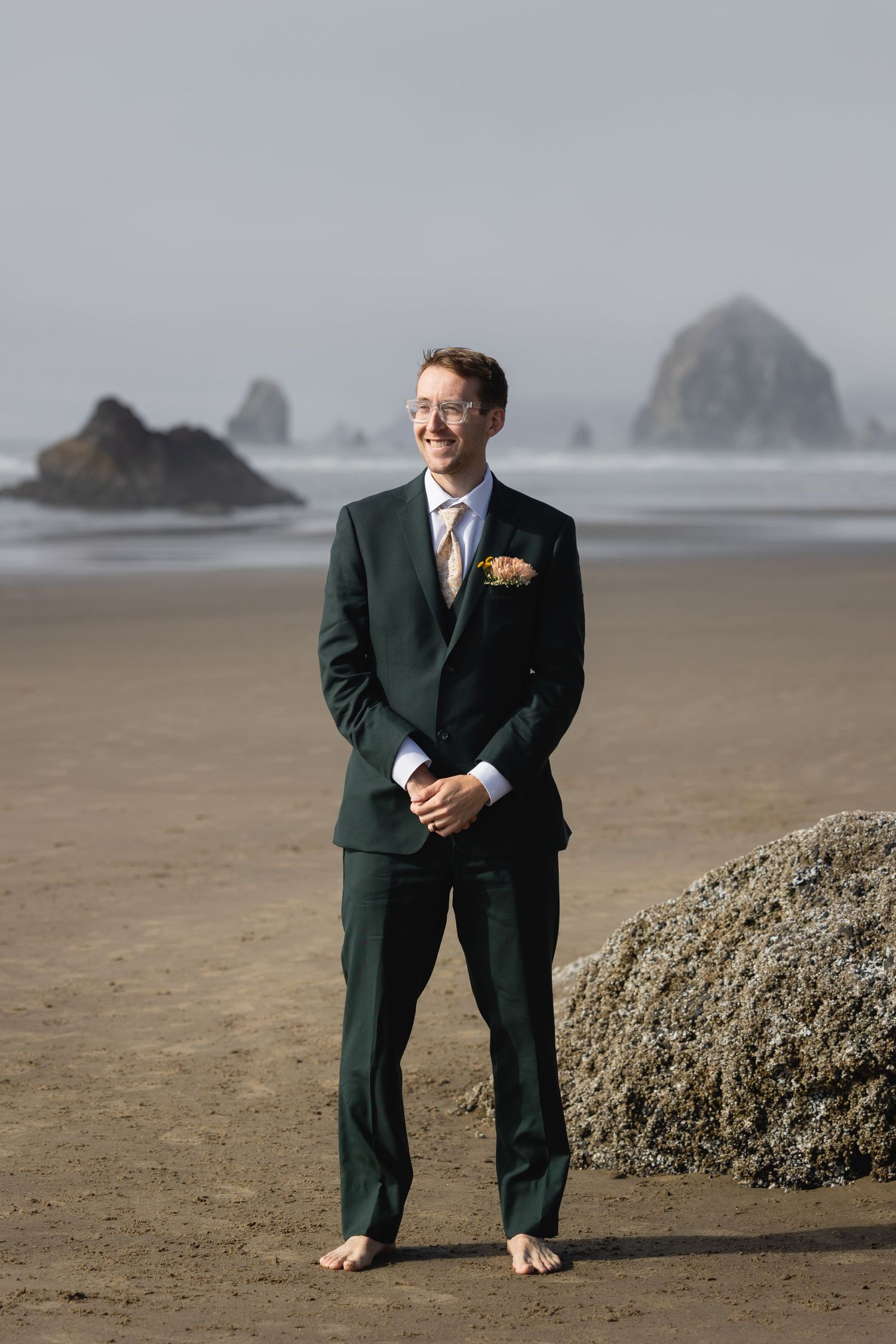 A person in a dark suit stands barefoot on a misty beach near rocky formations.