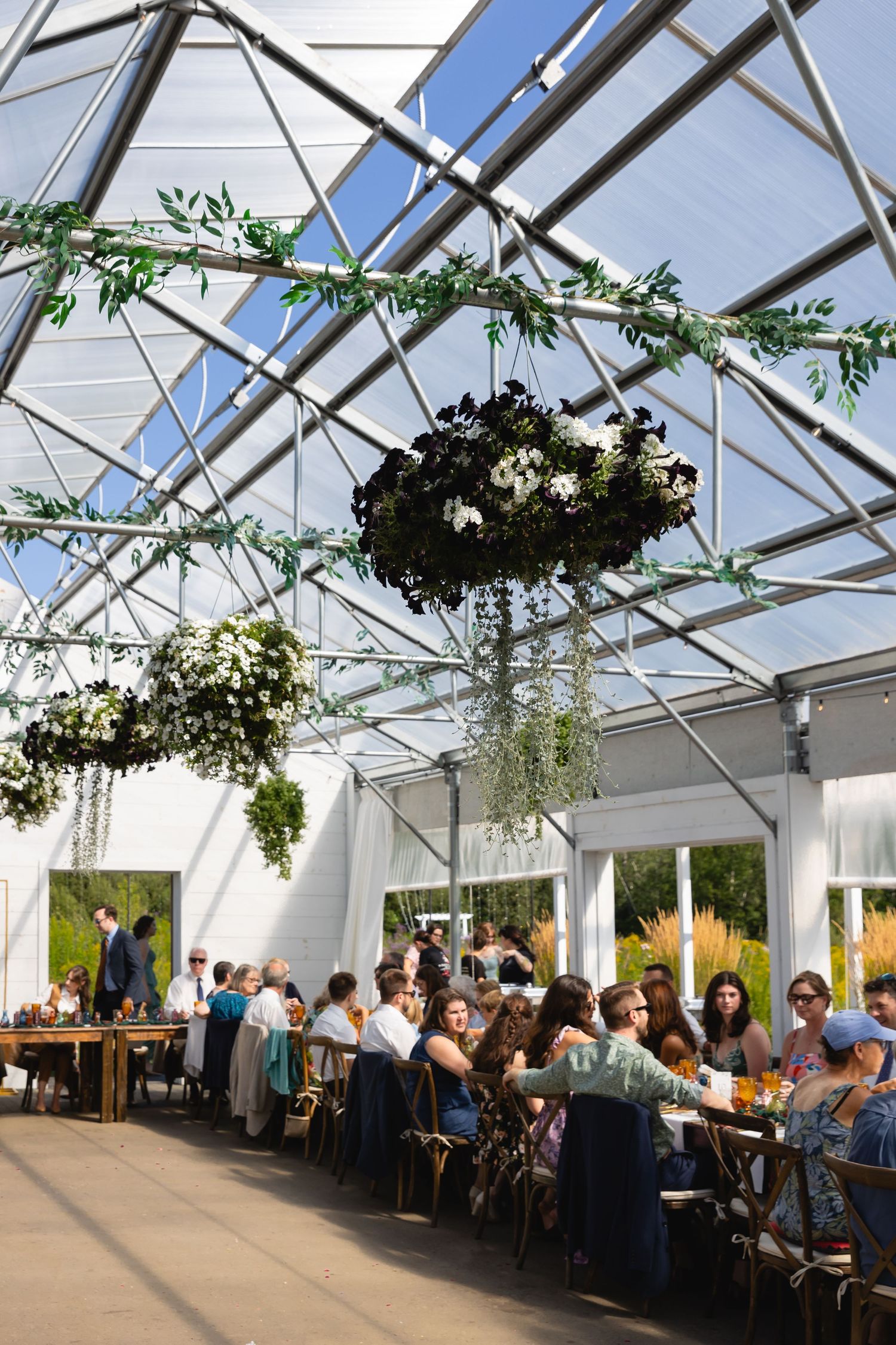 Wedding reception guests seated at long tables under a bright greenhouse-style tent with hanging floral arrangements.