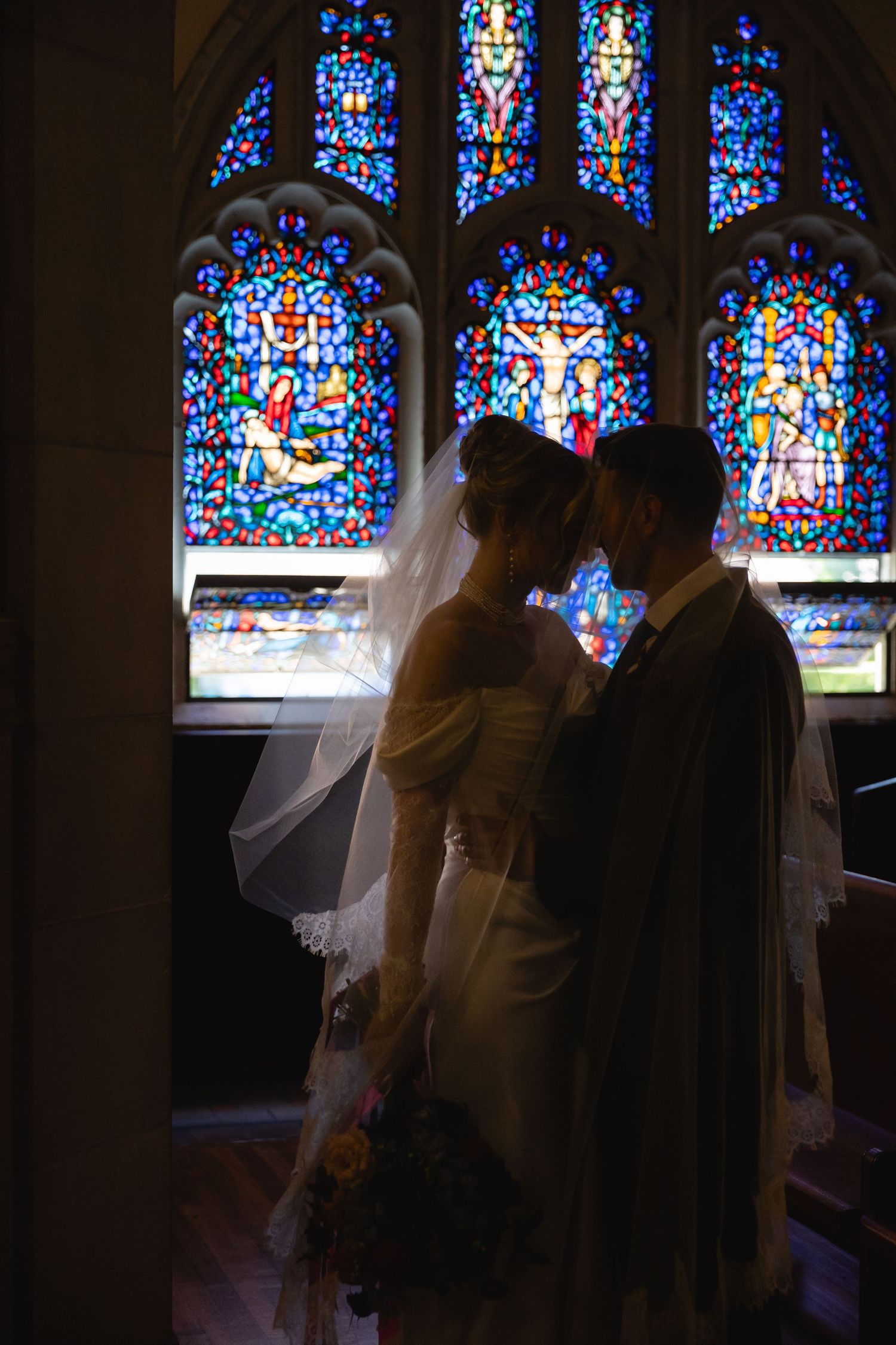 A silhouetted couple shares an intimate moment in front of colorful stained glass church windows.