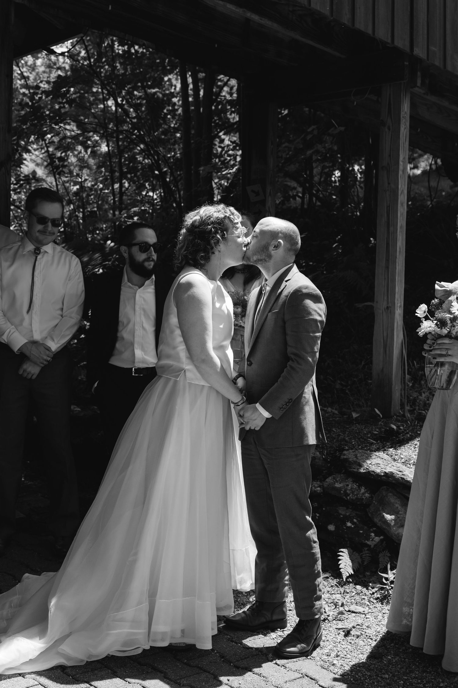 Wedding couple and guests gather under an outdoor pavilion during a sunny garden ceremony.