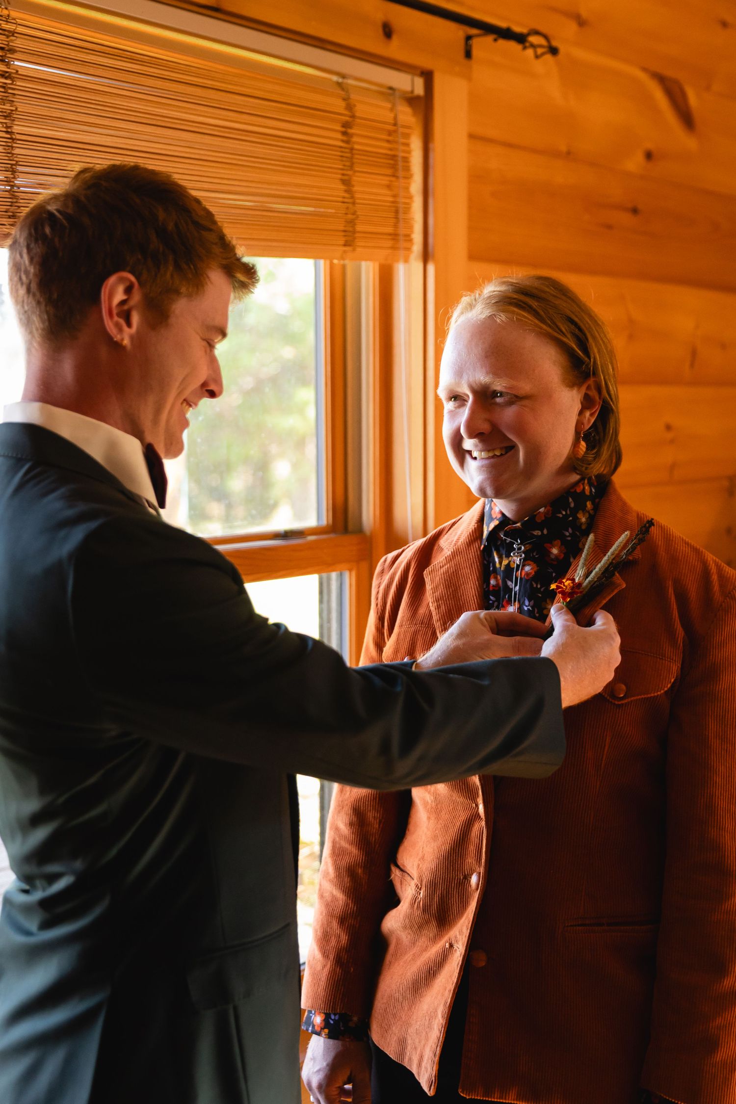 Two people in formal attire share a lighthearted moment while adjusting clothing in a wood-paneled room.