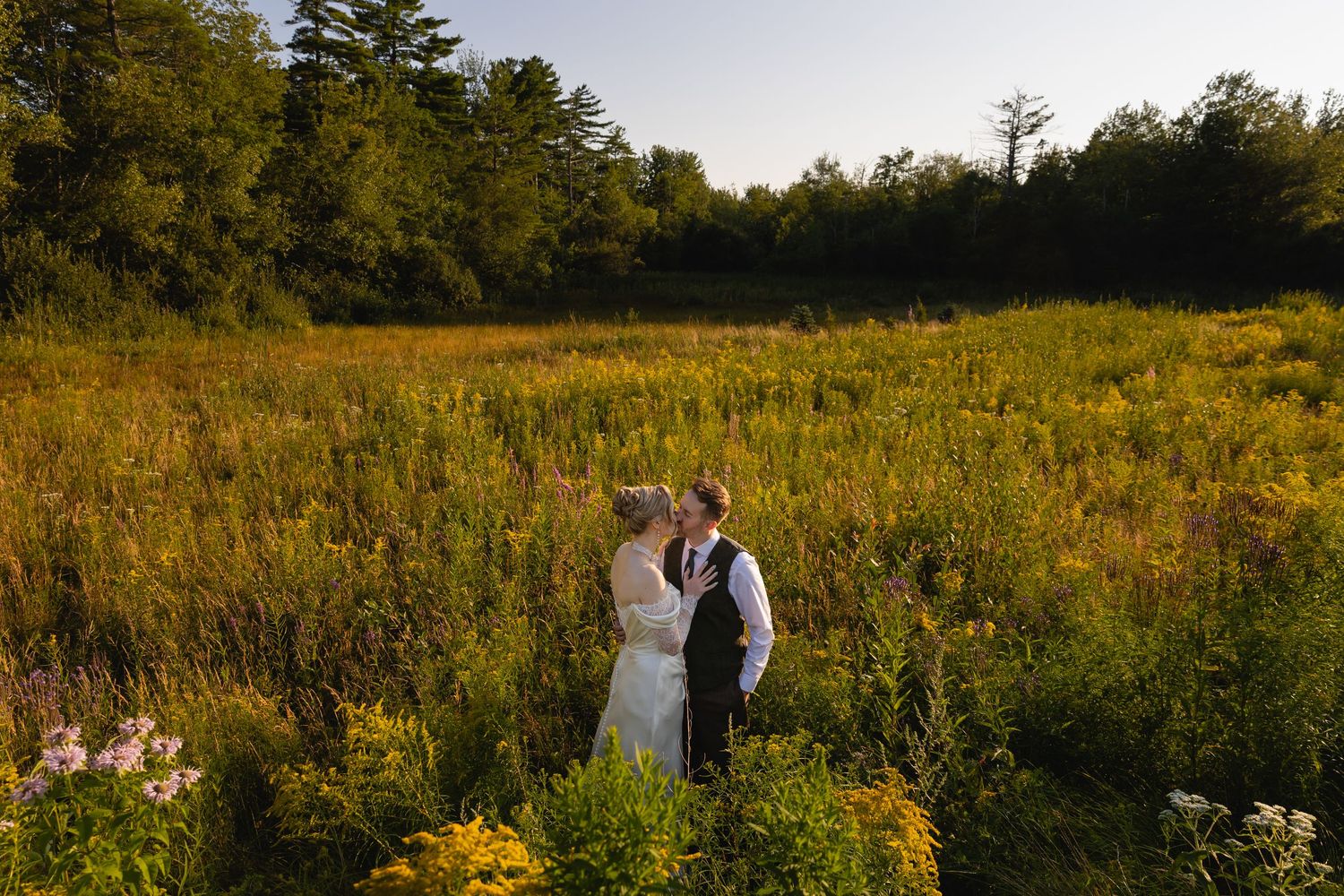 A couple shares a romantic moment in a golden meadow at sunset surrounded by wildflowers and tall grass.