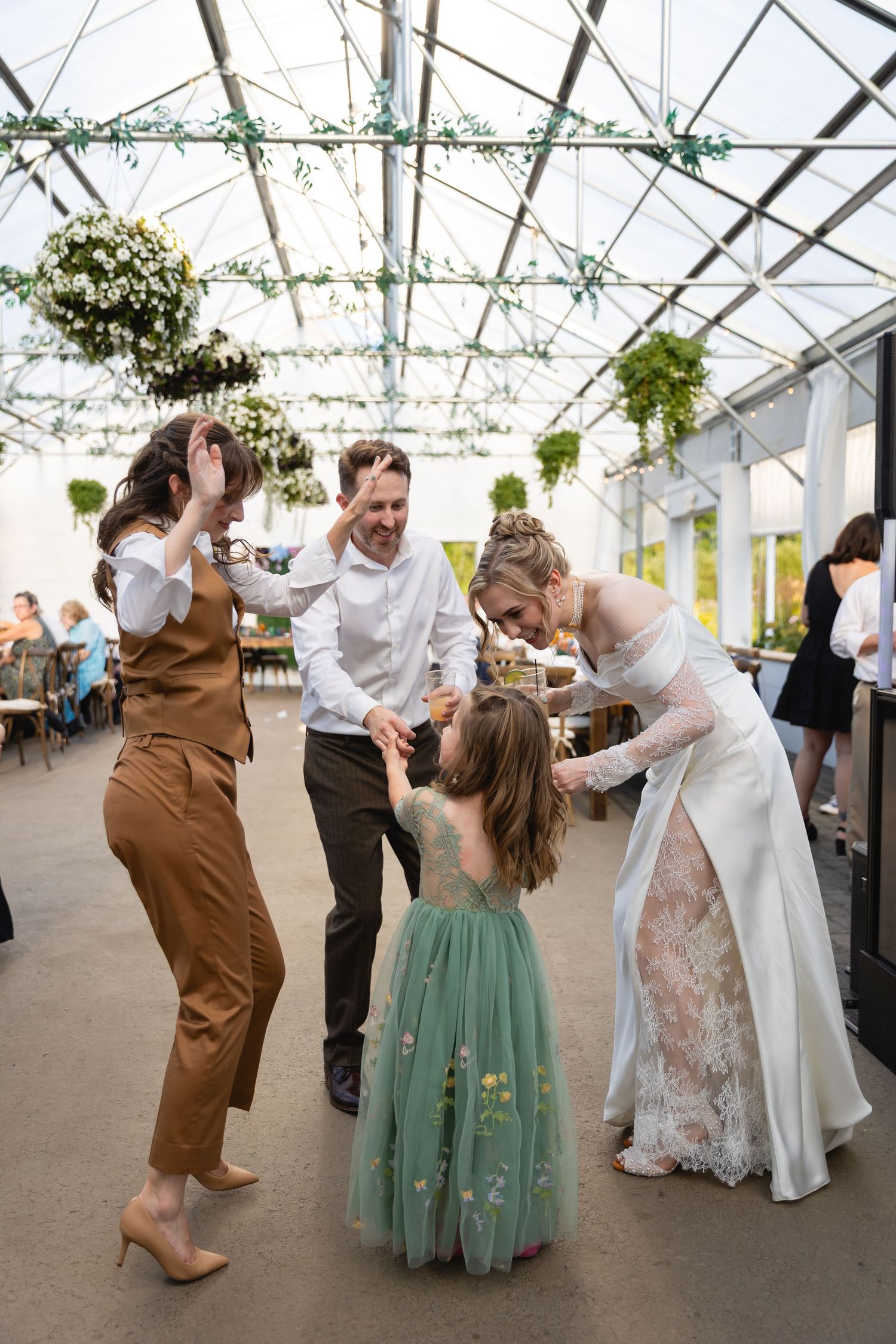 Wedding guests dance and twirl together under a greenhouse tent with hanging greenery decorations at a celebration.