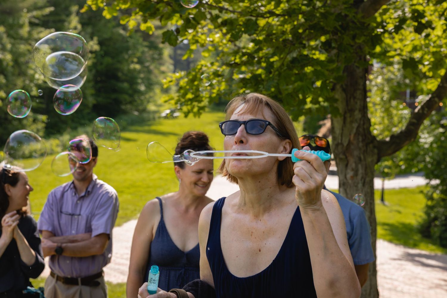 People blowing bubbles together in a sunny park setting with trees and green grass in the background.