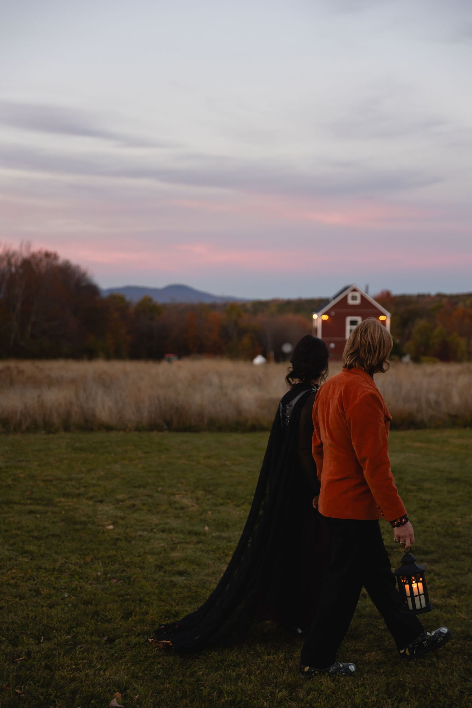 Two people in autumn attire walk across a grassy field at sunset with mountains and a red barn in the distance.
