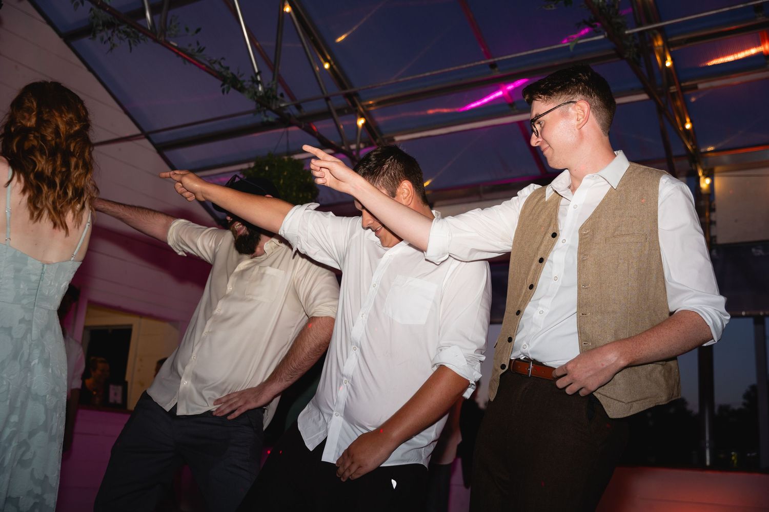 Party guests dance energetically under colorful lighting at an indoor celebration event.