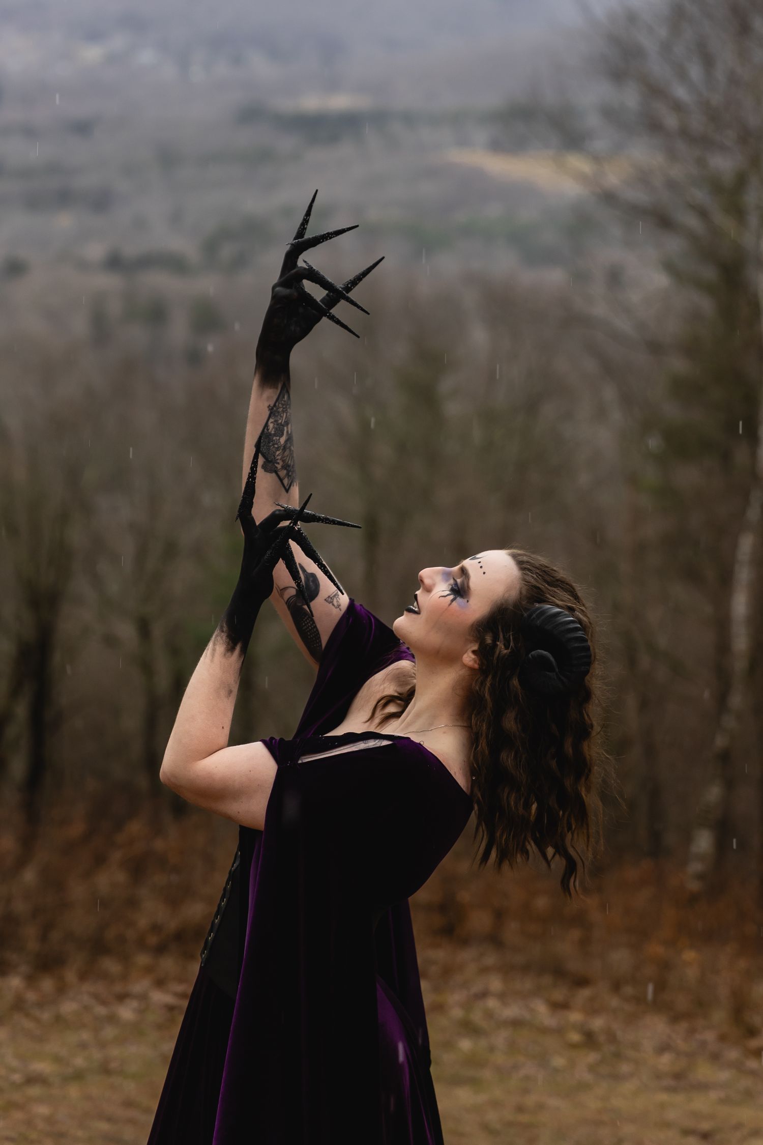 Woman in purple velvet dress with black spiked gloves reaching skyward in misty outdoor setting.