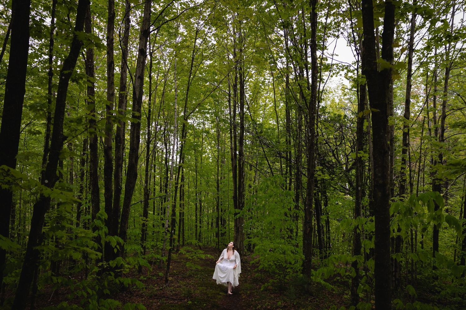 Woman in white dress walks through a lush green forest path surrounded by tall trees.