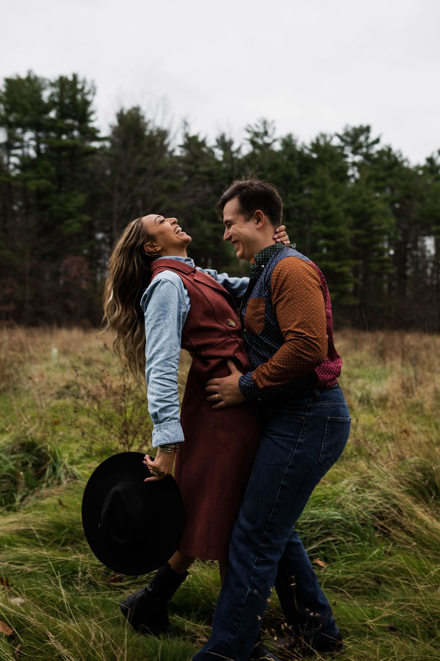A playful autumn photoshoot in a field with tall grass and pine trees in the background.