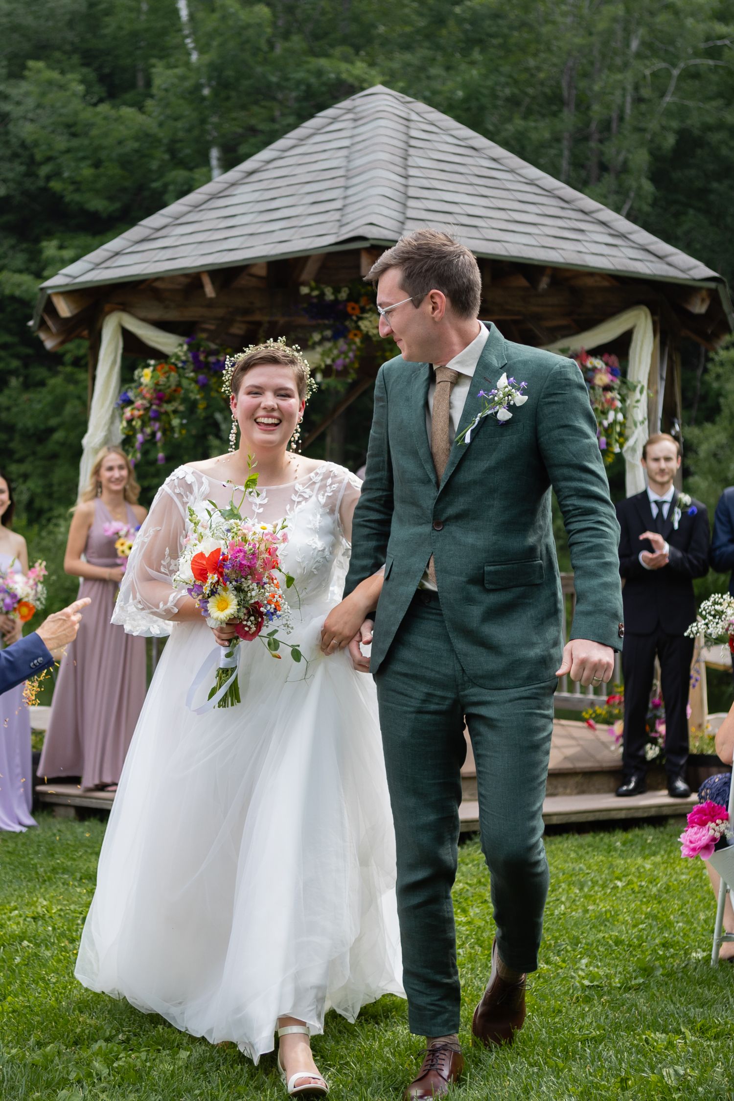 A couple celebrates their wedding day in front of a rustic gazebo surrounded by greenery.