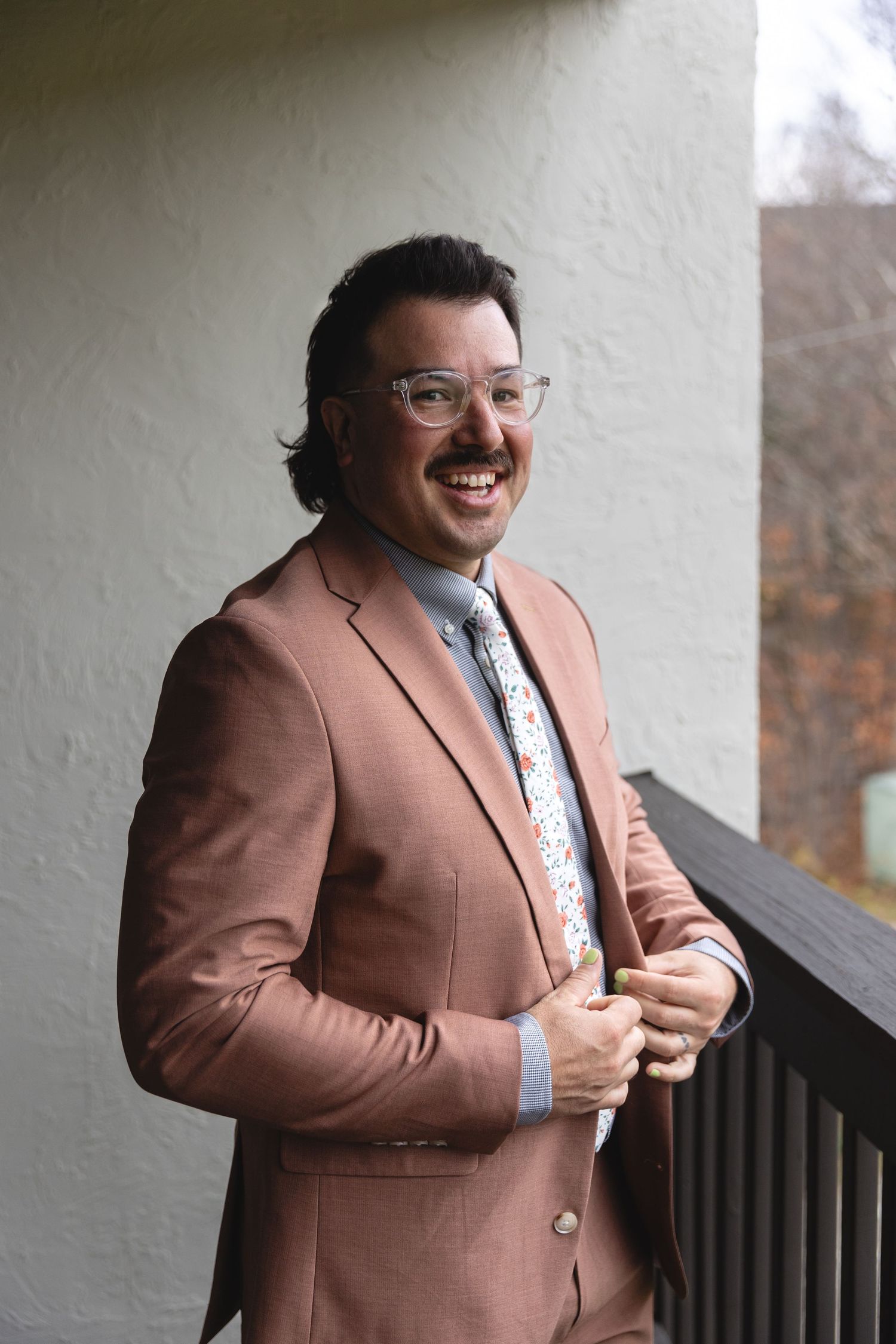 Person in a brown blazer and floral tie smiling on a porch.