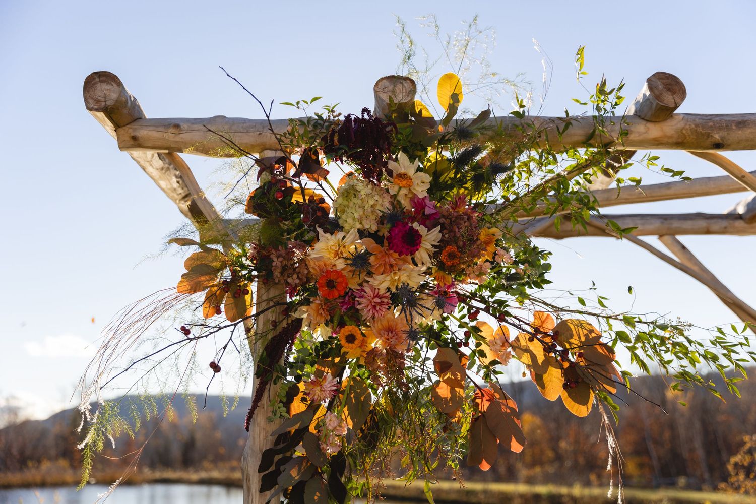 Vibrant floral arrangement adorning a wooden wedding arch with autumn foliage against a blue sky.