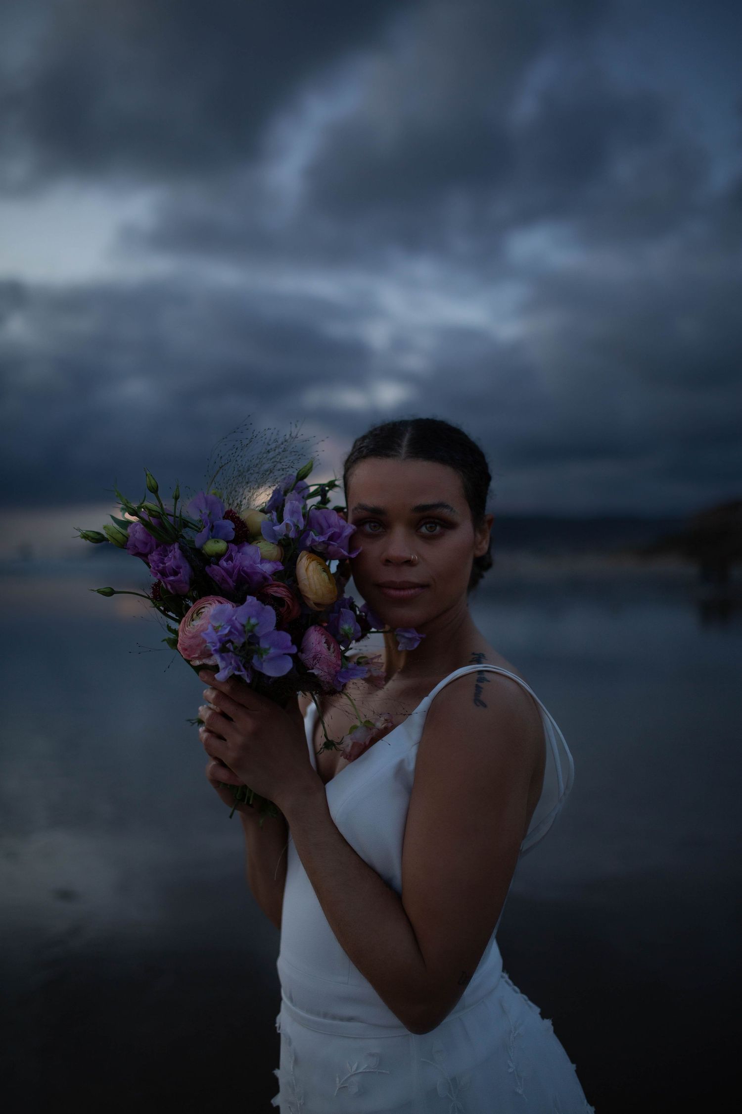 A person in a white wedding dress holds a vibrant purple bouquet against stormy beach scenery.
