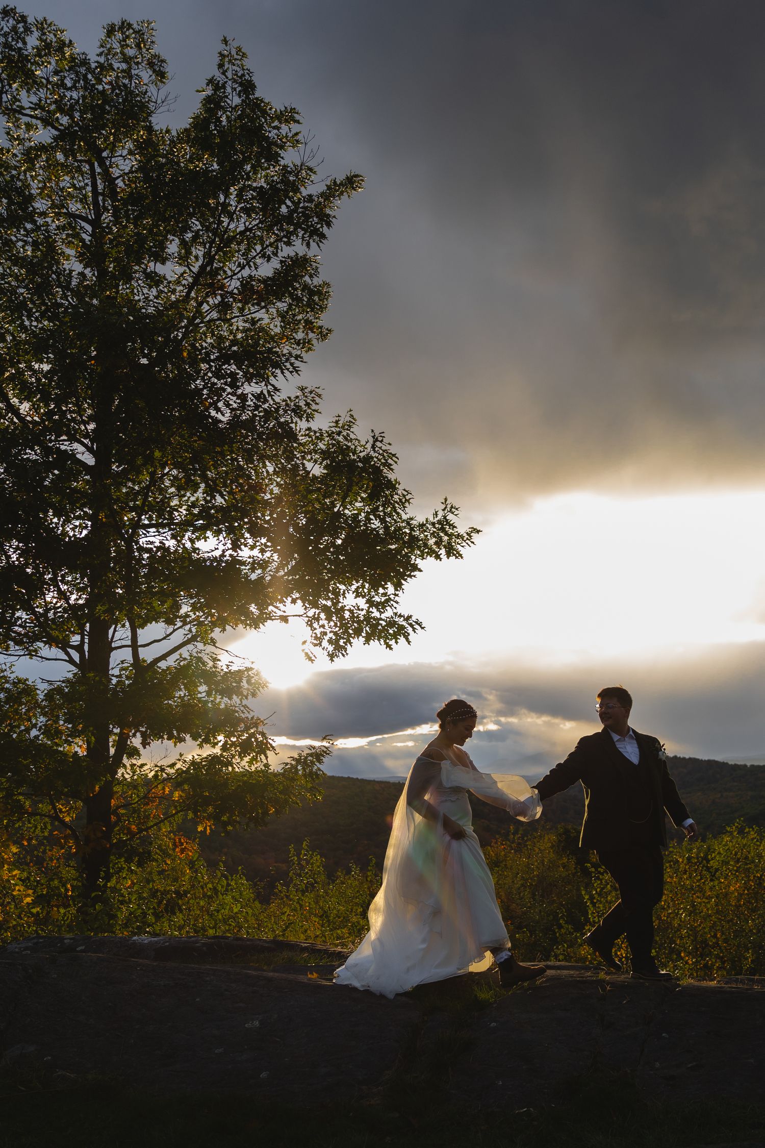 Newlyweds walk hand in hand on mountain overlook with dramatic sunset behind them.
