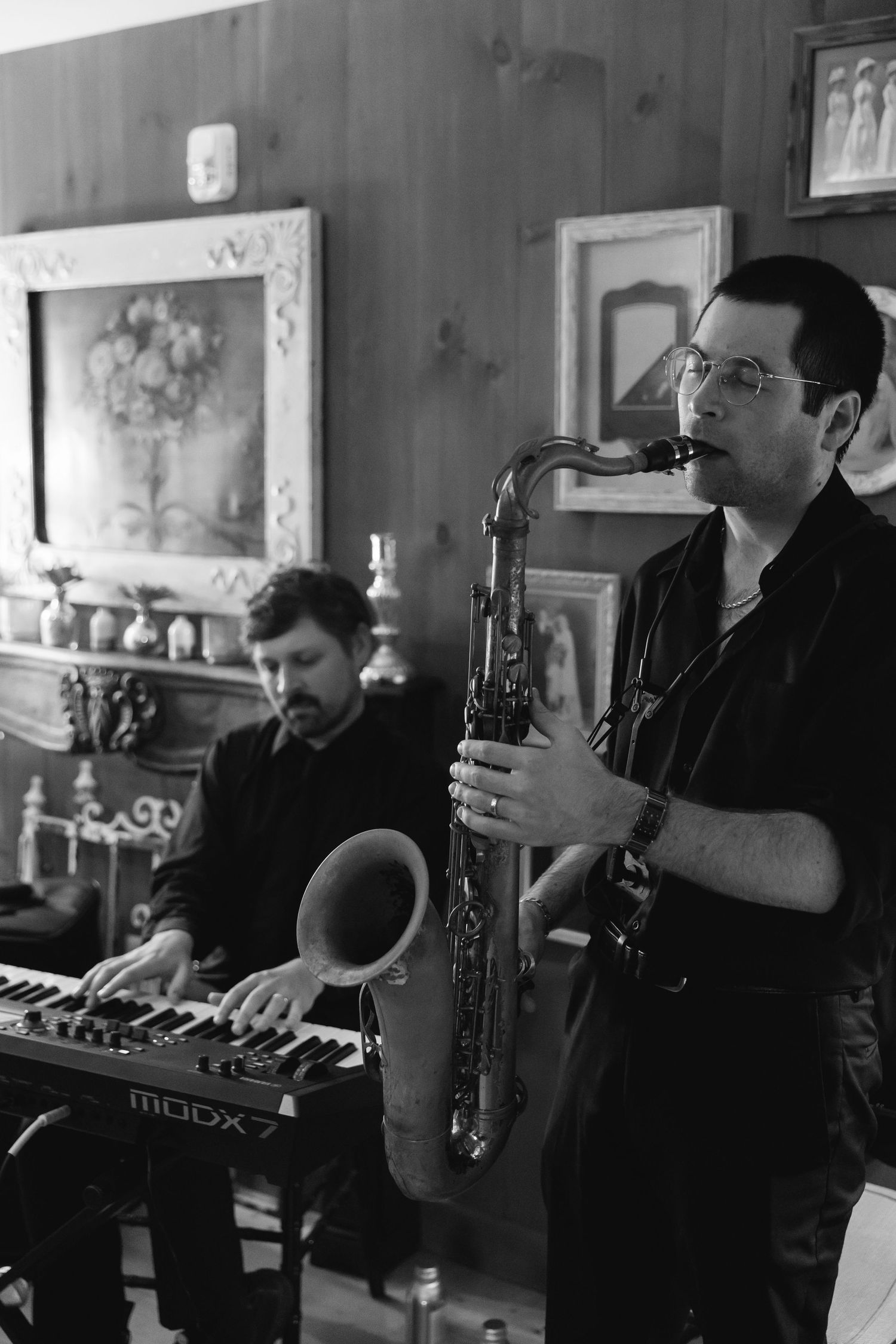 Two jazz musicians perform together, one playing a saxophone and another on a keyboard in a black and white photo.