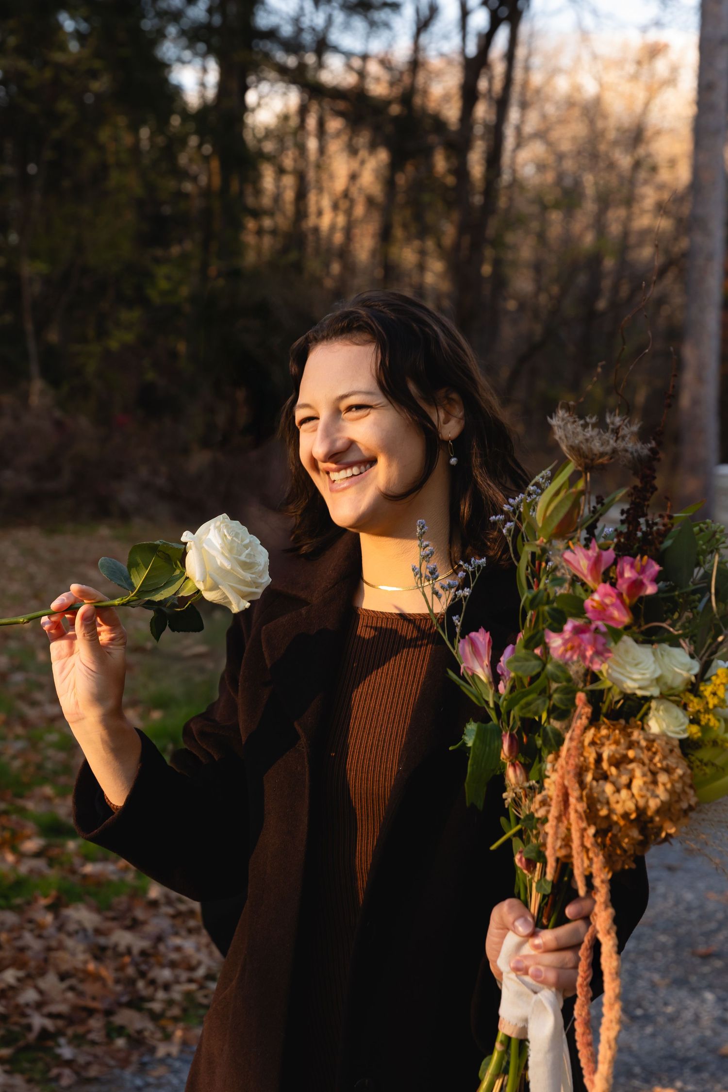 A person in a brown sweater holds a bouquet of mixed flowers while smiling outdoors during golden hour.