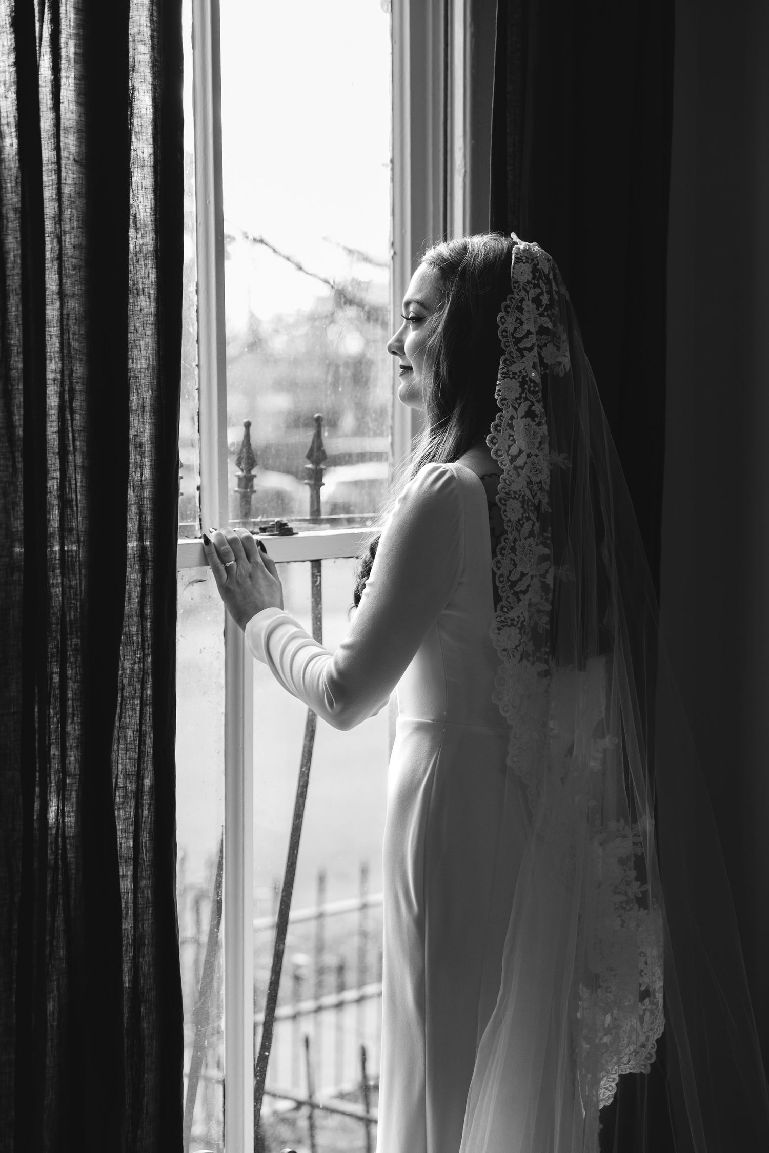 A bride in a flowing white gown and veil looks pensively out a tall window in black and white photography.