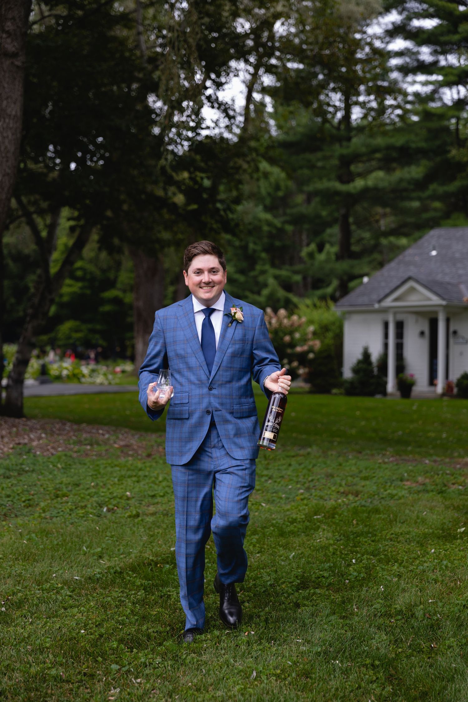 Someone in a blue plaid suit walks across a green lawn while holding a drink bottle.