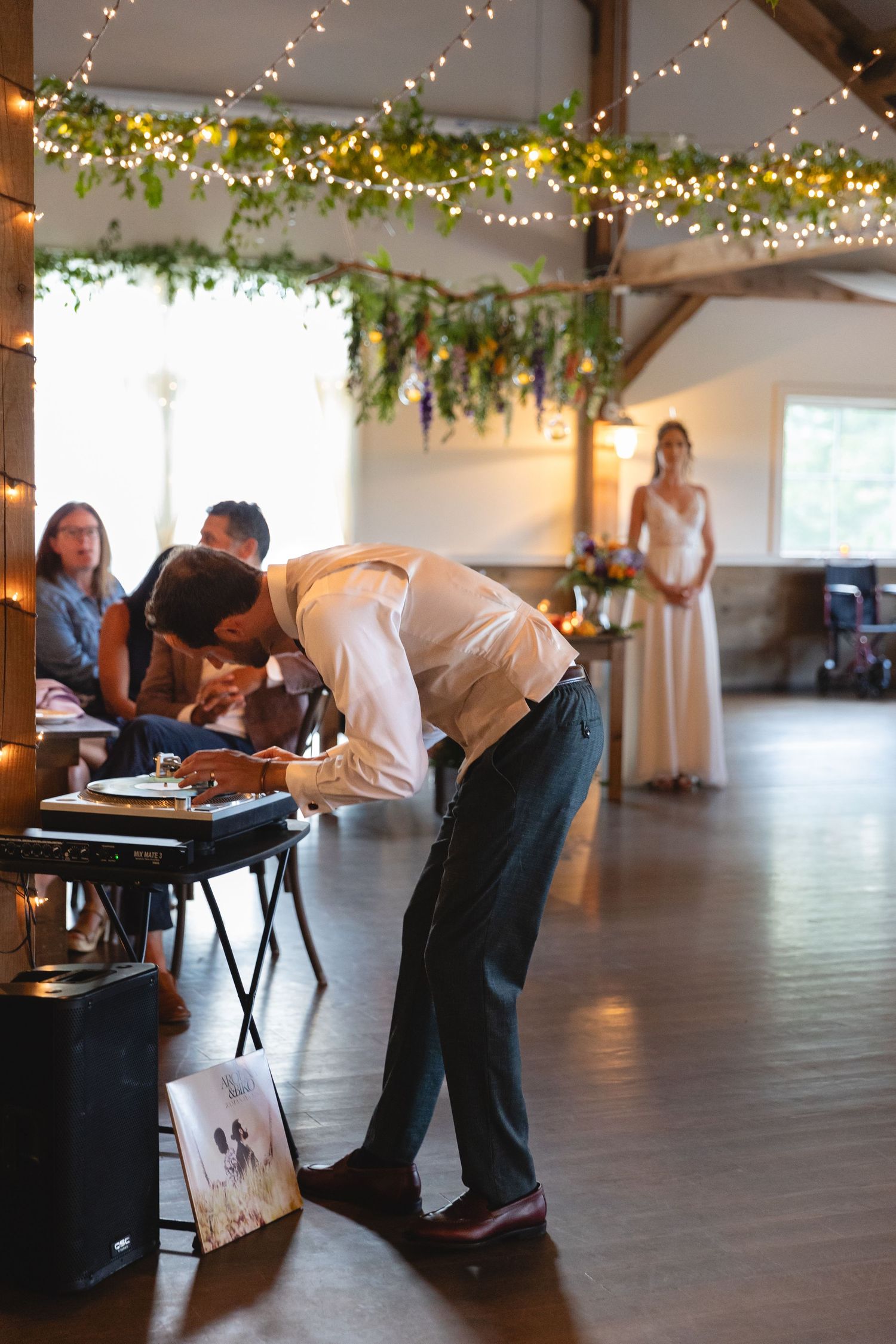 Event DJ working turntables in rustic venue with string lights.